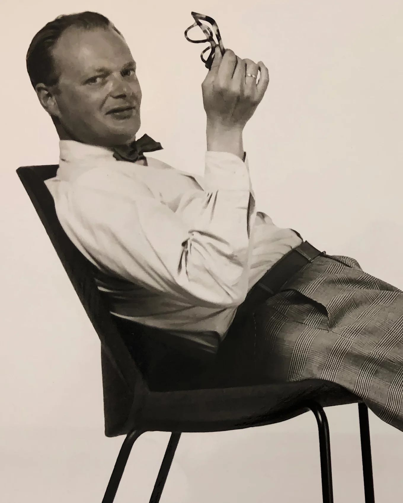 Homme en nœud papillon et pantalon à carreaux se penchant en arrière sur une chaise, tenant des lunettes et souriant à l'appareil.