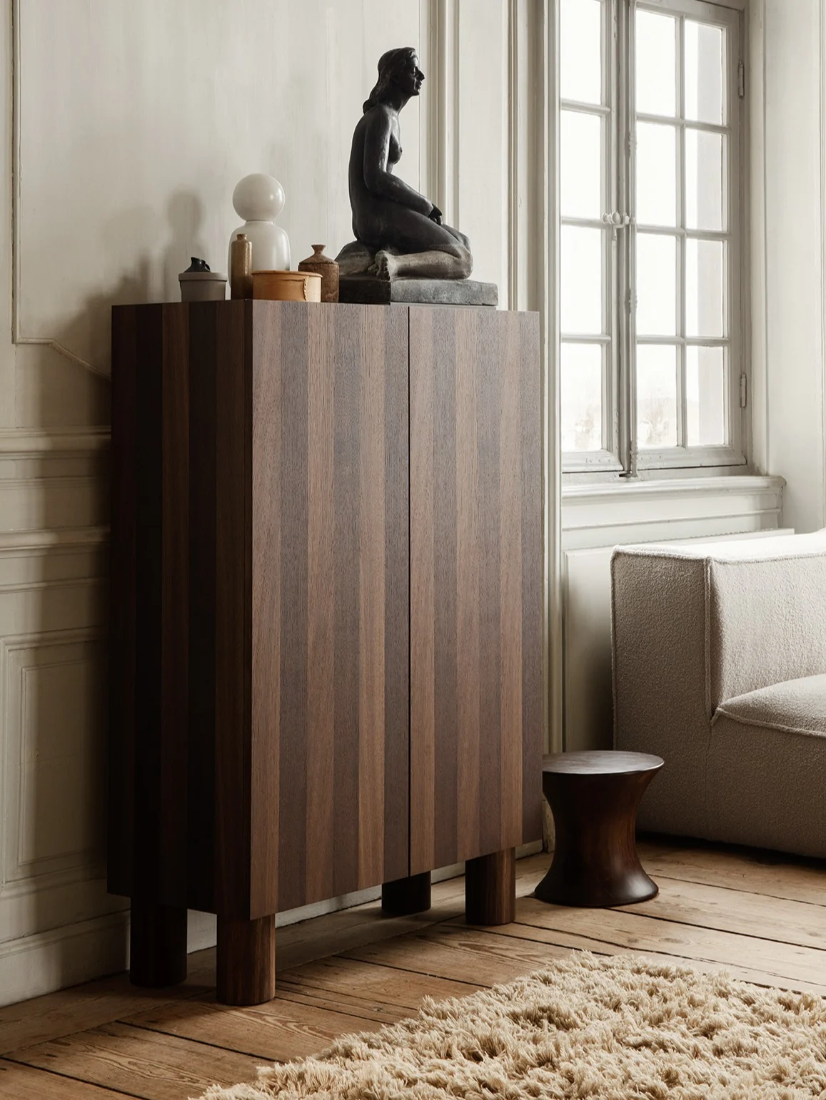 Meuble en bois foncé à rayures verticales, surmonté d'une sculpture assise, d'une lampe blanche et de décors, à côté d'un canapé et d'un tabouret sombre.
