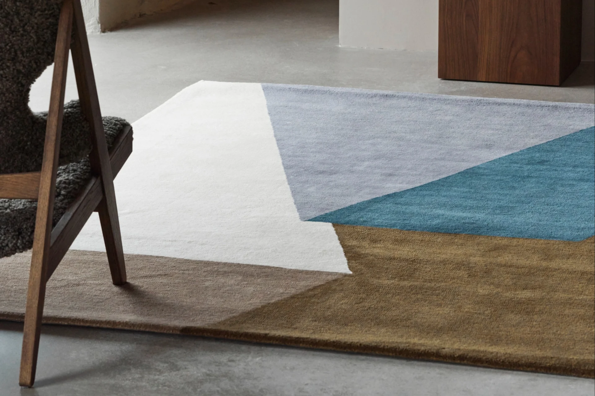 Un tapis géométrique blanc, bleu clair, turquoise et marron sur un sol en béton, avec une chaise en bois et un vase bleu.