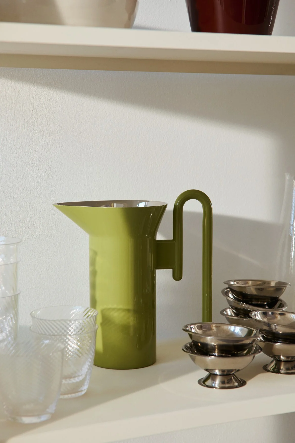 Une carafe vert clair dotée d'une anse unique en forme de U, des verres transparents texturés et de petits bols argentés posés sur une étagère.