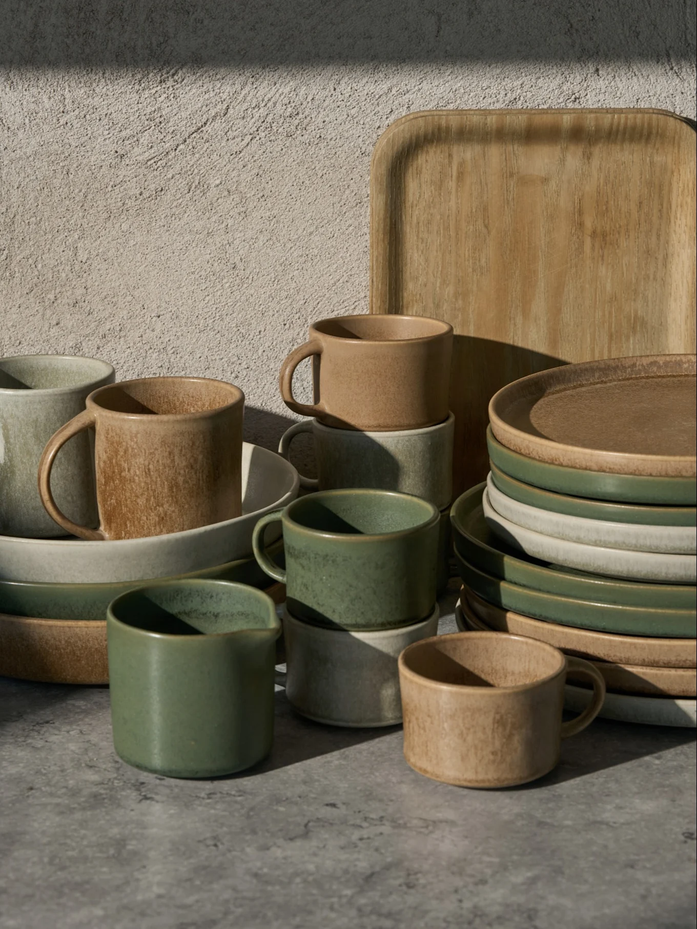 Collection de tasses, bols et assiettes en céramique verts, beiges et gris clair, avec un plateau en bois, sur une surface grise.