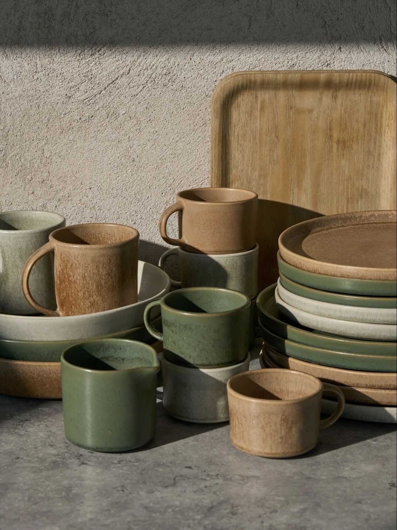 Collection de tasses, bols et assiettes en céramique verts, beiges et gris clair, avec un plateau en bois, sur une surface grise.