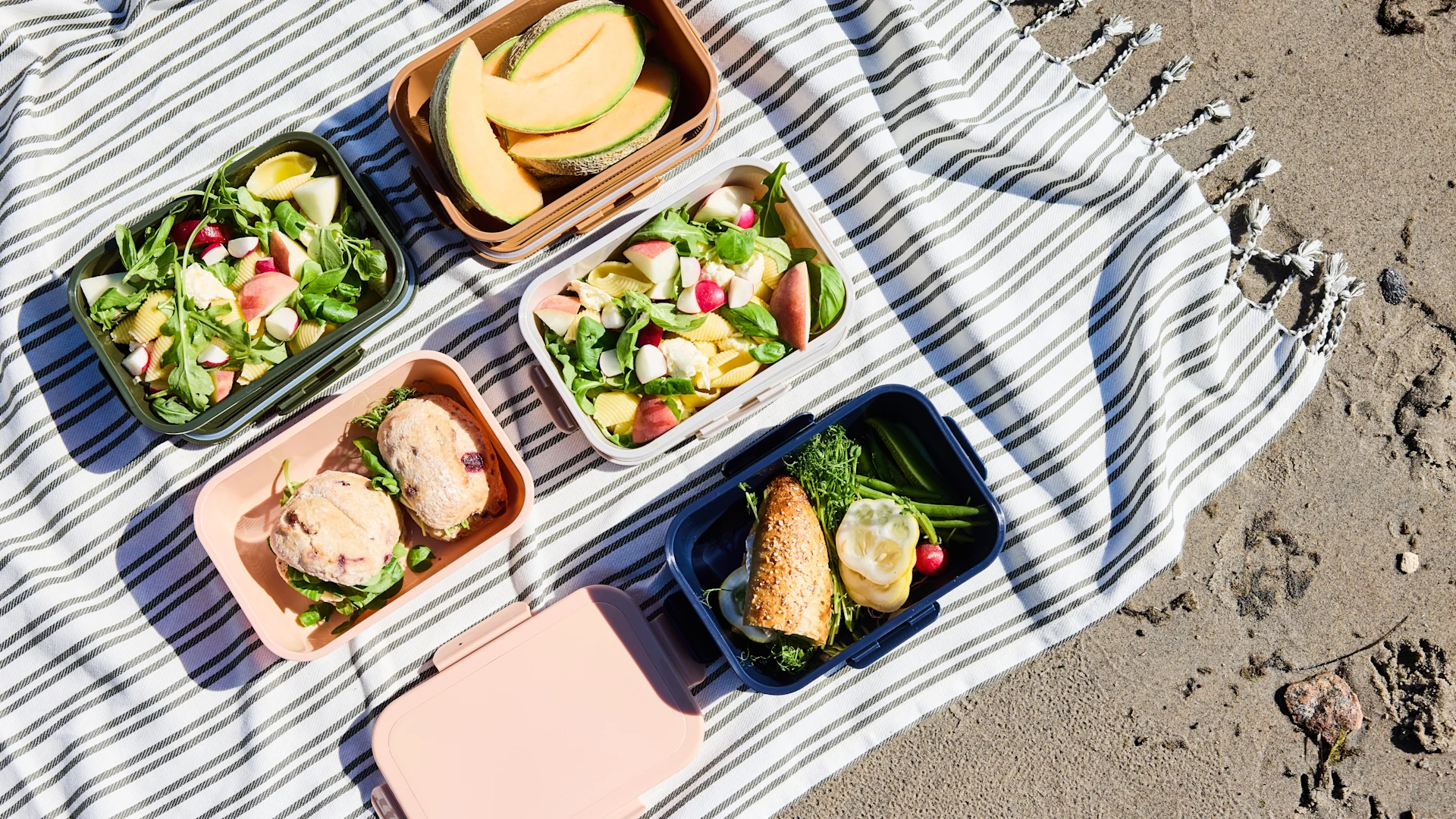 Pique-nique sur une couverture rayée, posée sur le sable, avec plusieurs boîtes à lunch contenant des fruits, une salade de pâtes et des sandwichs.