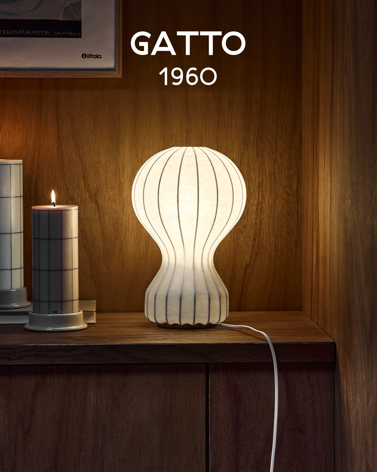Une lampe blanche striée lumineuse en forme de sablier sur une étagère en bois à côté d'une bougie allumée. Texte : GATTO 1960.