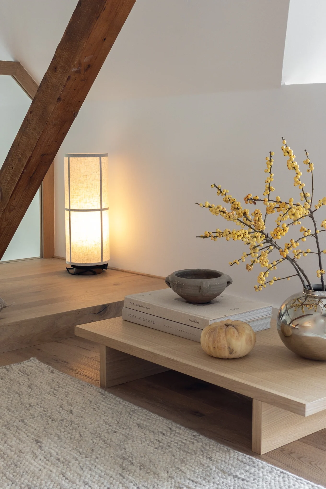 Coin salon mansardé douillet avec lampe haute, poutre en bois, table basse, baies jaunes, livres et petite citrouille.
