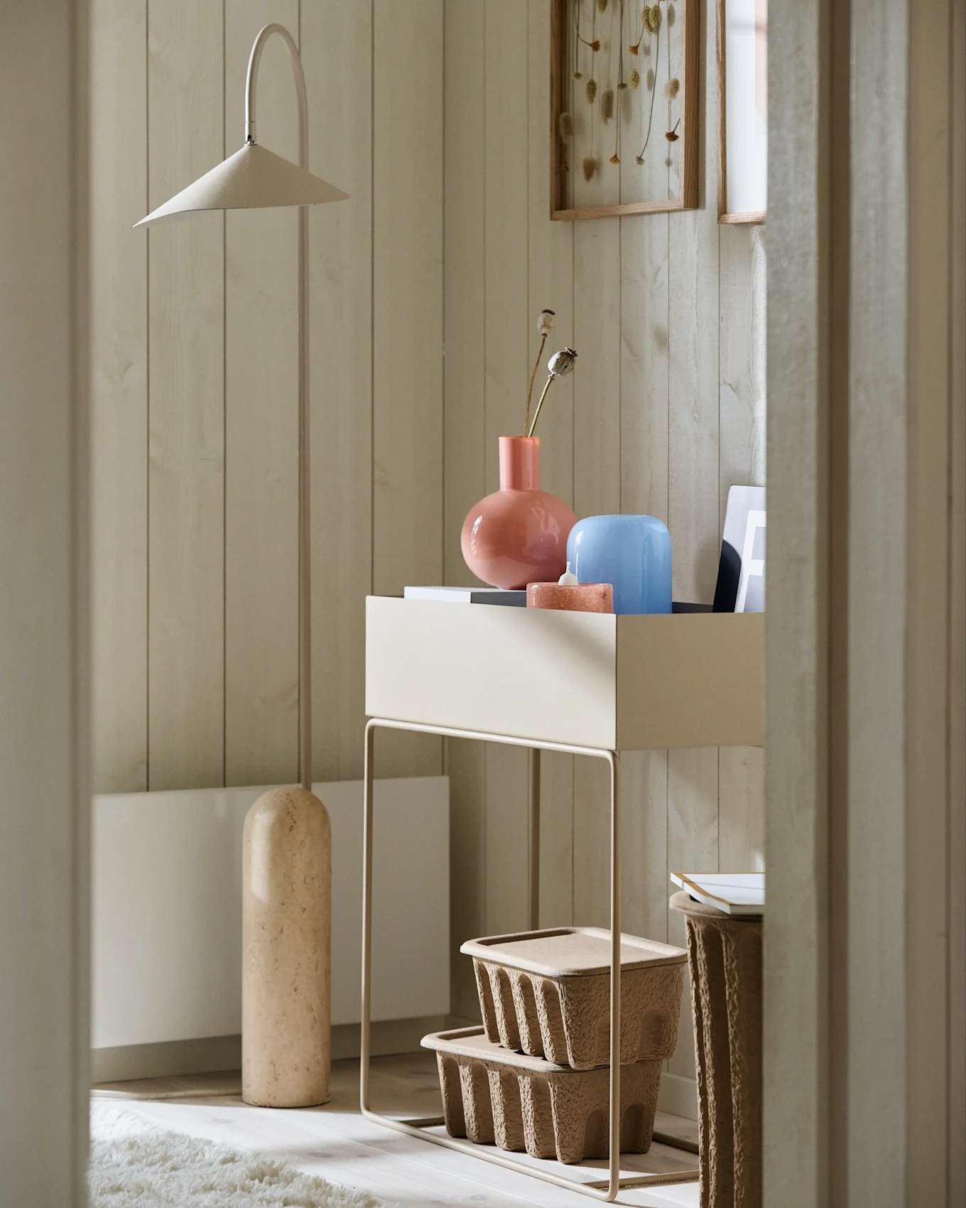 Coin minimaliste avec un grand lampadaire beige, une console avec des vases roses et bleus, et des boîtes de rangement marron texturées.