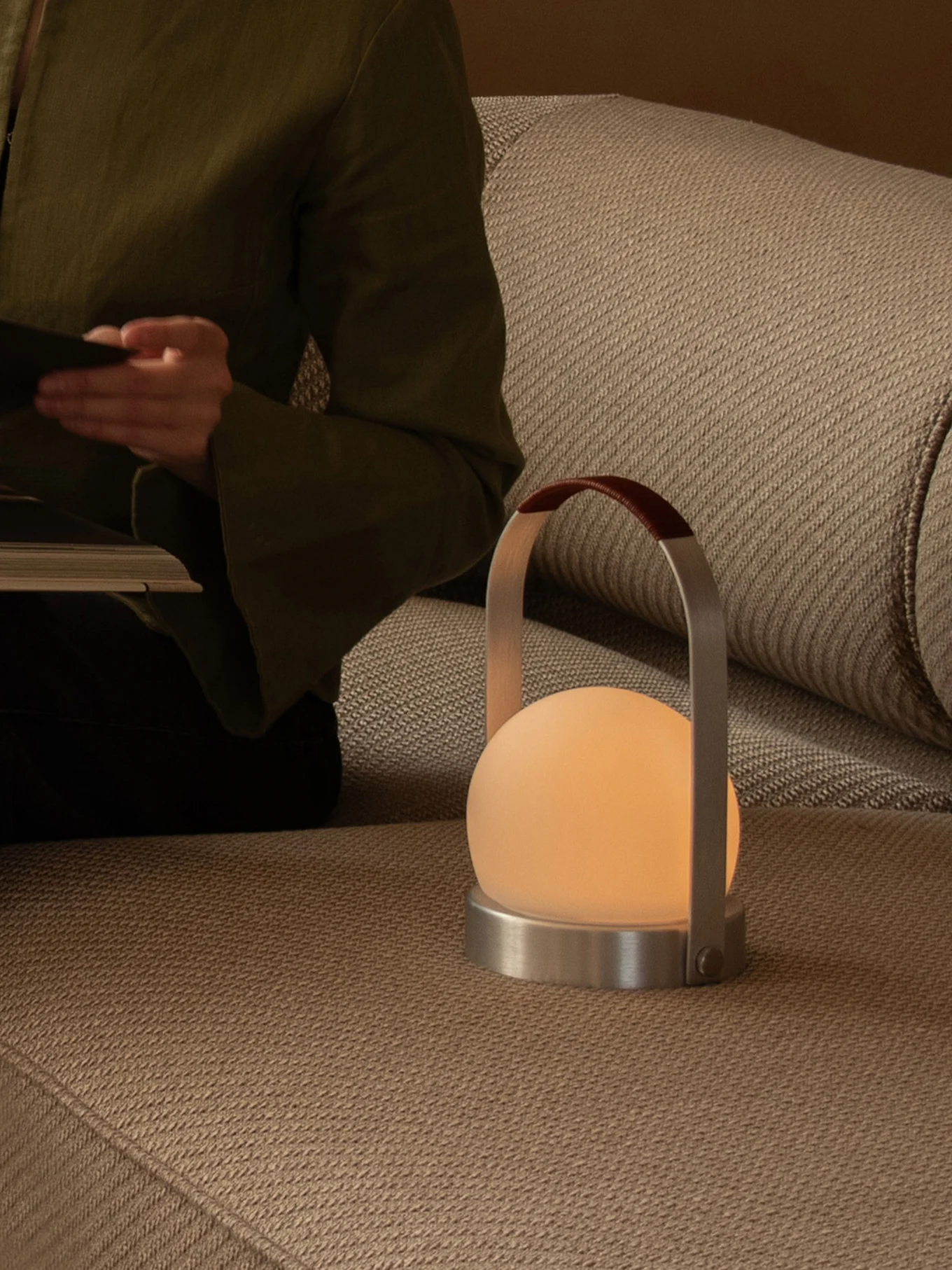 Personne lisant sur un canapé, à côté d'une lampe portable lumineuse avec globe dépoli, base métallique et anse en cuir.