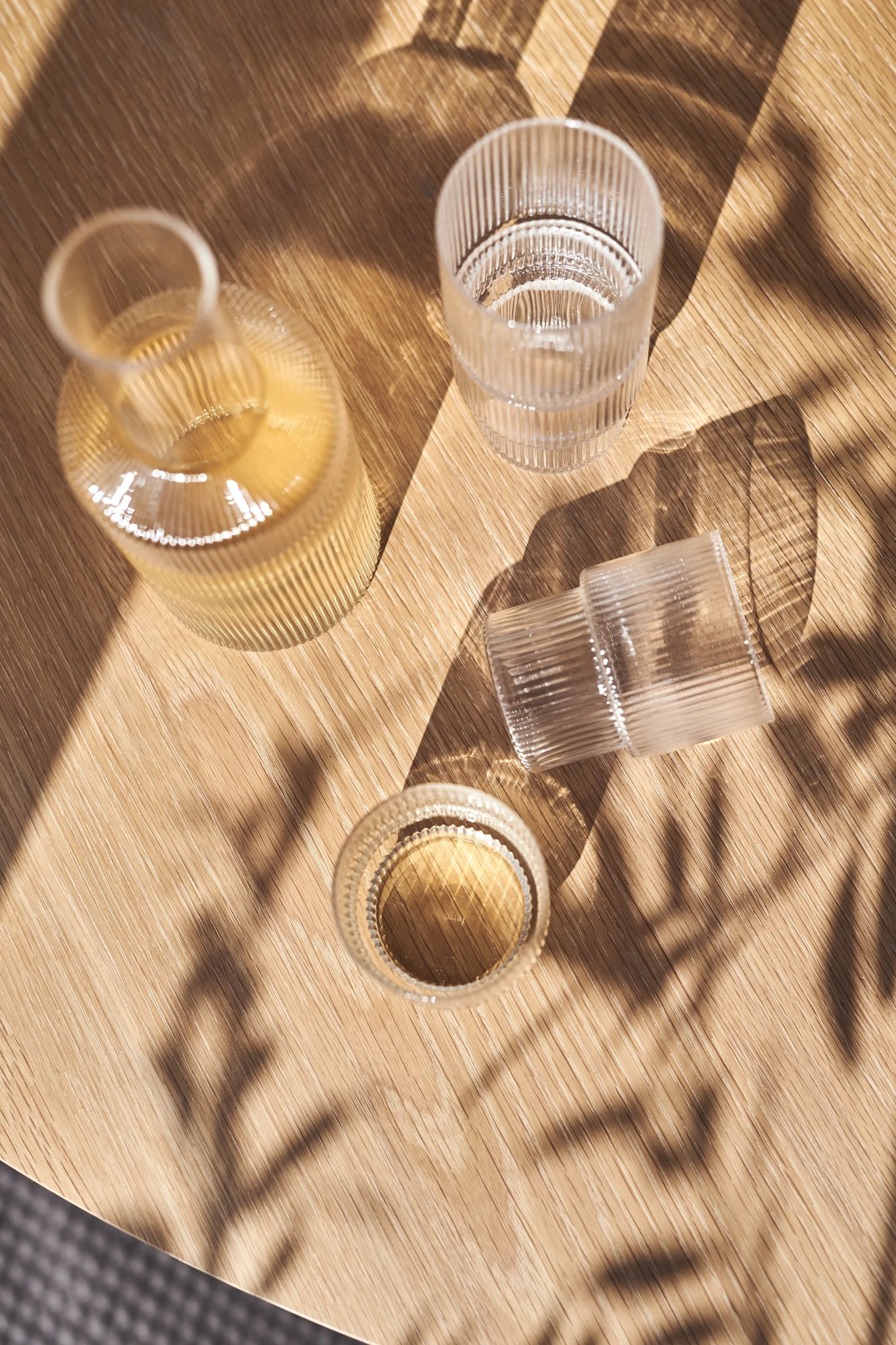 Une bouteille en verre côtelée et deux verres côtelés, un avec du liquide, sur une table en bois ensoleillée avec des ombres de feuilles.