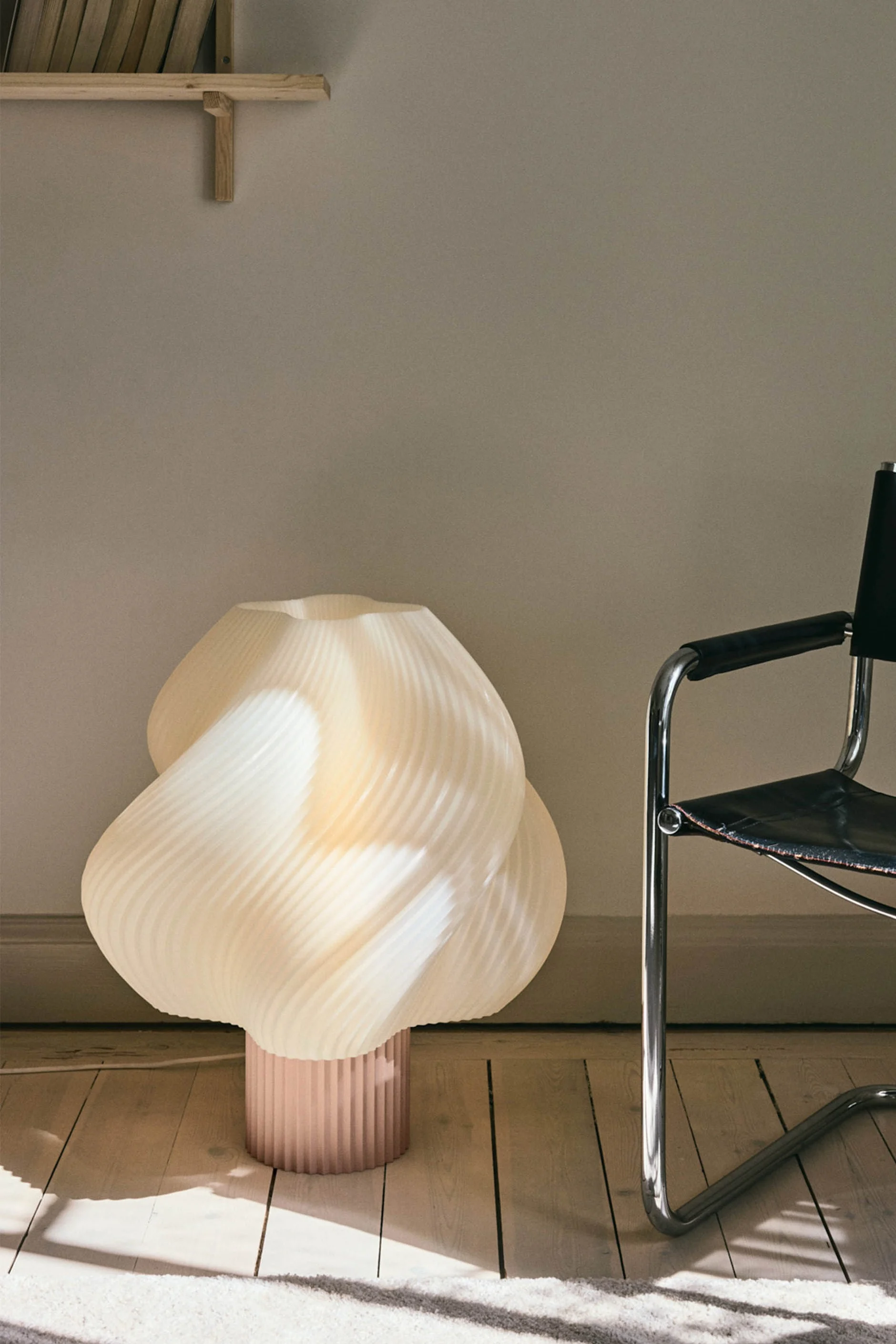 Un lampadaire sculptural de couleur crème, aux plis élégants, doté d'un pied rose, trône sur un parquet, à côté d'une chaise en cuir noir et chrome.