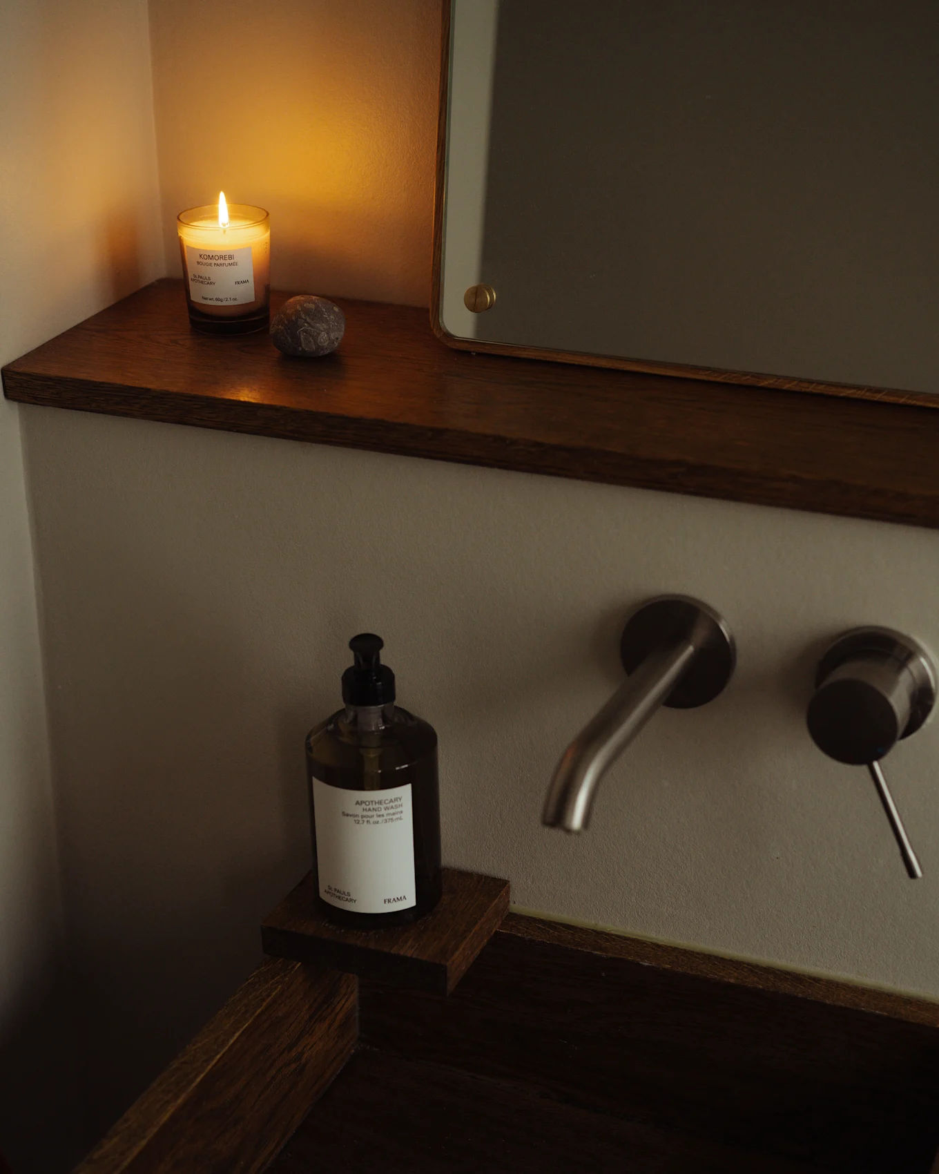 Salle de bain chaleureuse avec bougie allumée, flacon de savon pour les mains et robinet en métal brossé sur lavabo en bois foncé.