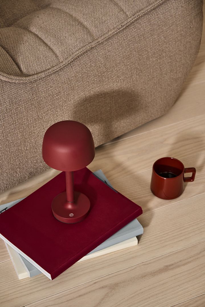 Le rouge foncé est l'une des tendances de la décoration intérieure pour 2025. Ici, la lampe de table Halo sans fil de couleur bordeaux est posée sur une pile de livres, à côté de la tasse Taper rouge, toutes deux de Scandi Living.