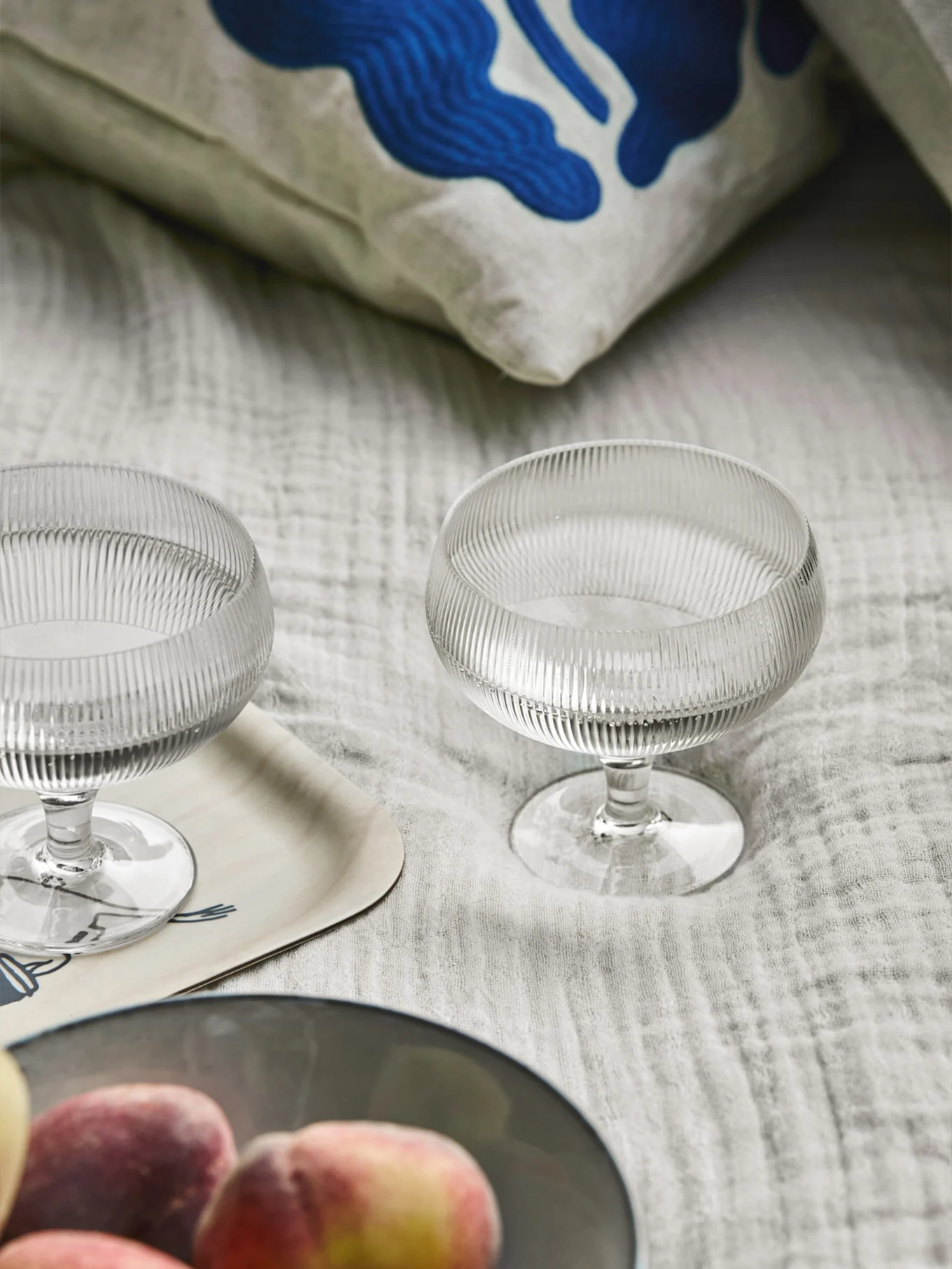 Deux coupes à dessert en verre côtelé, un oreiller avec un motif abstrait bleu, un plateau en bois et des pêches sur un tissu clair.