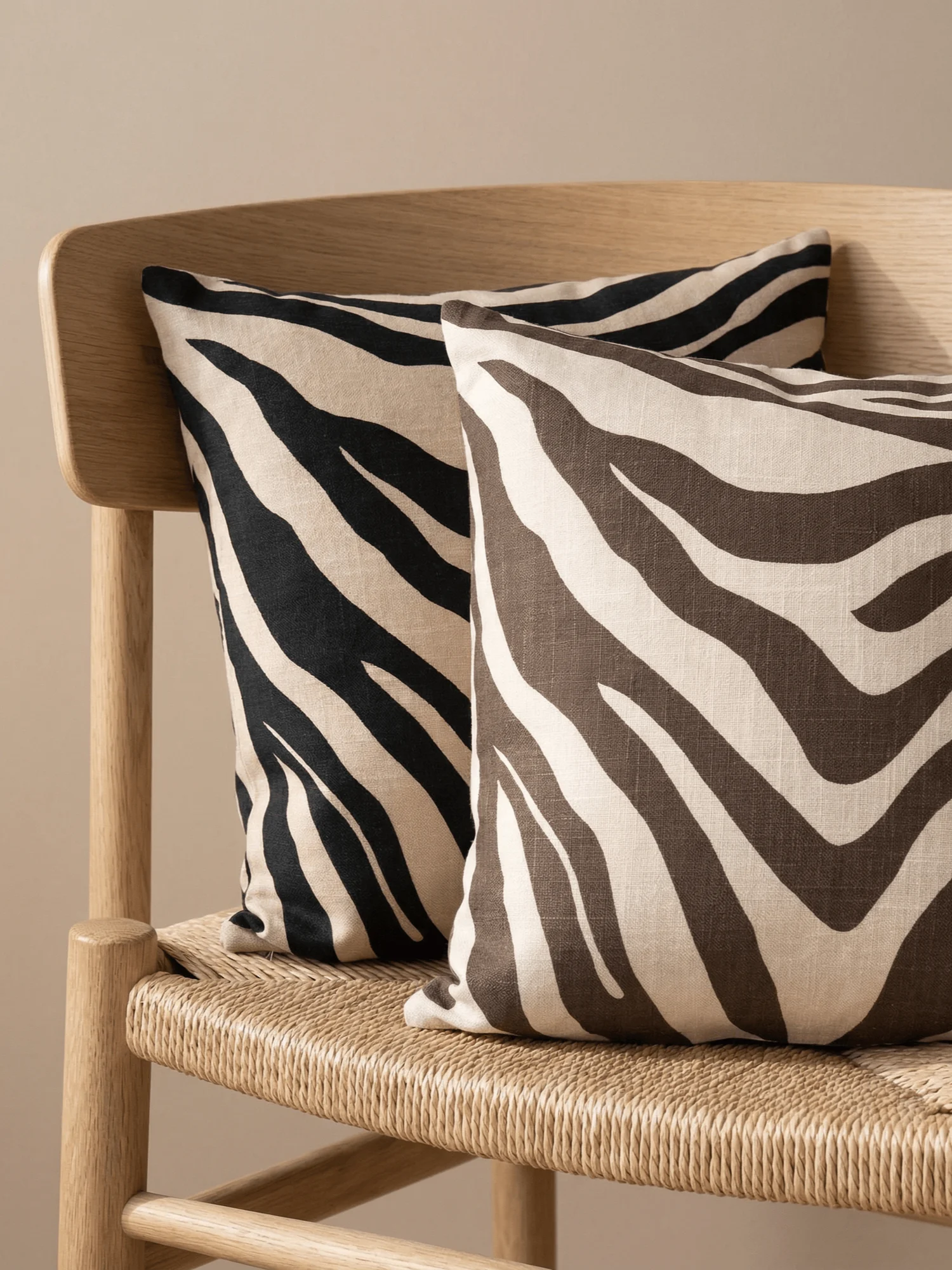 Deux coussins décoratifs à motif zèbre, l'un noir et crème, l'autre marron et crème, posés sur une chaise en bois tressé.