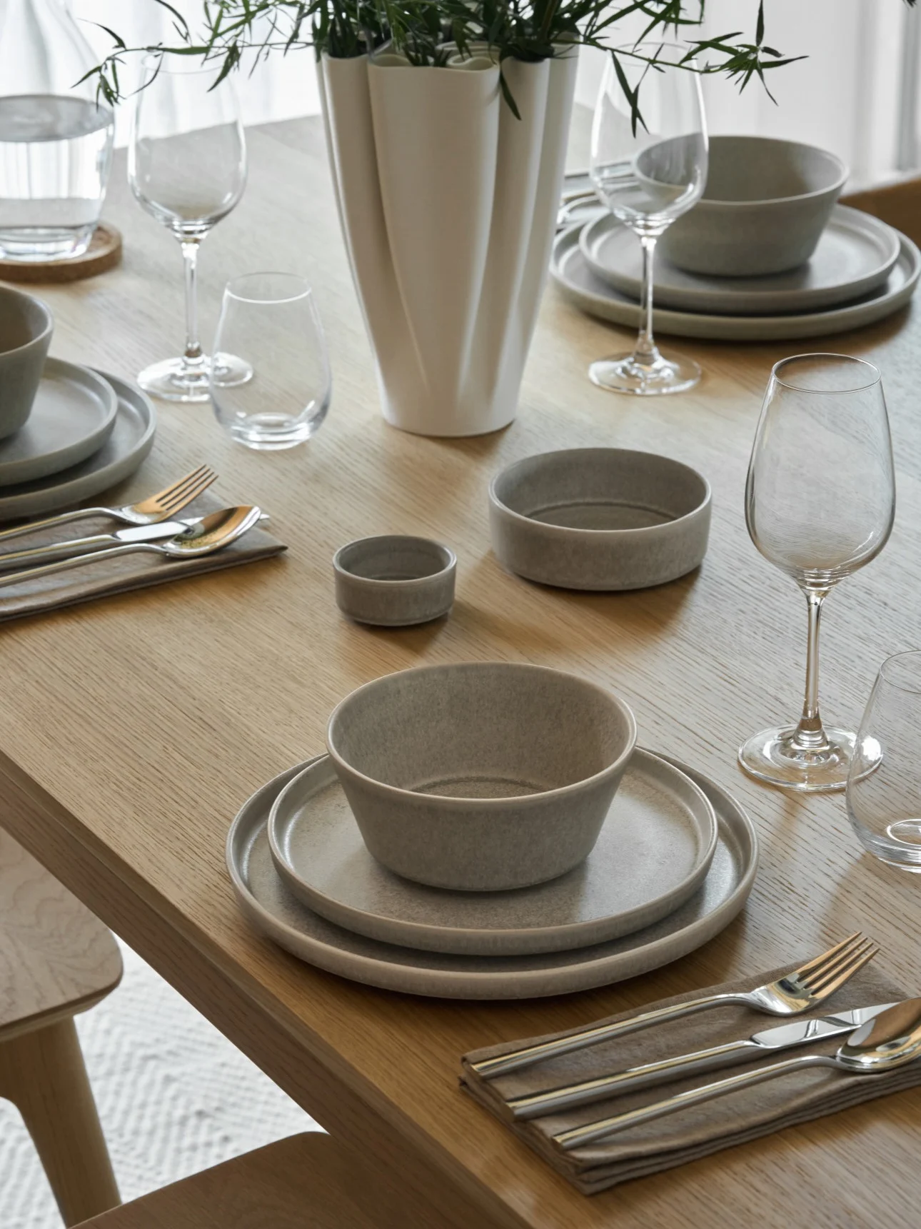 Une table à manger en bois clair dressée avec de la vaisselle en céramique grise, des couverts en argent, des verres transparents et un vase blanc avec du feuillage vert.