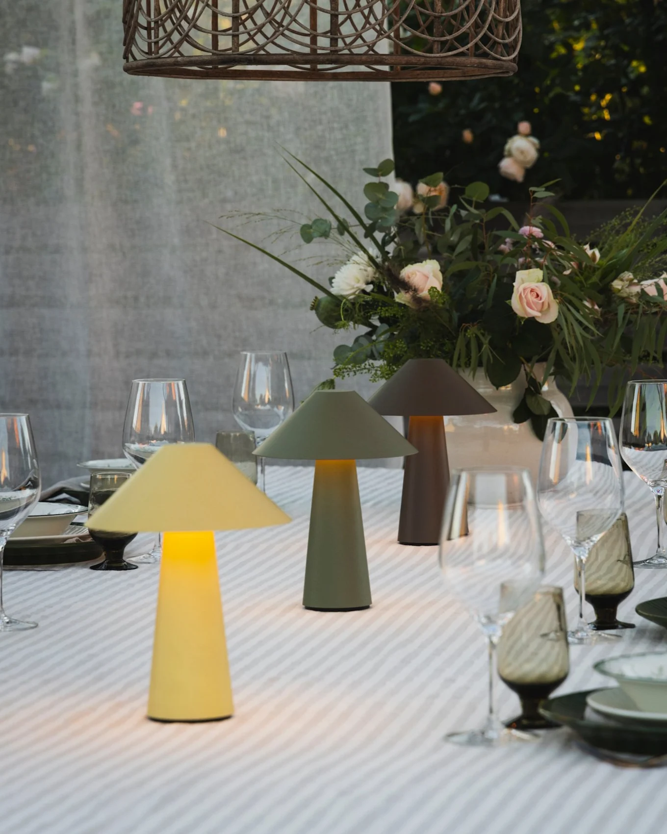 Table à manger extérieure avec trois lampes champignon éclairées, un centre de table floral et une suspension en osier.