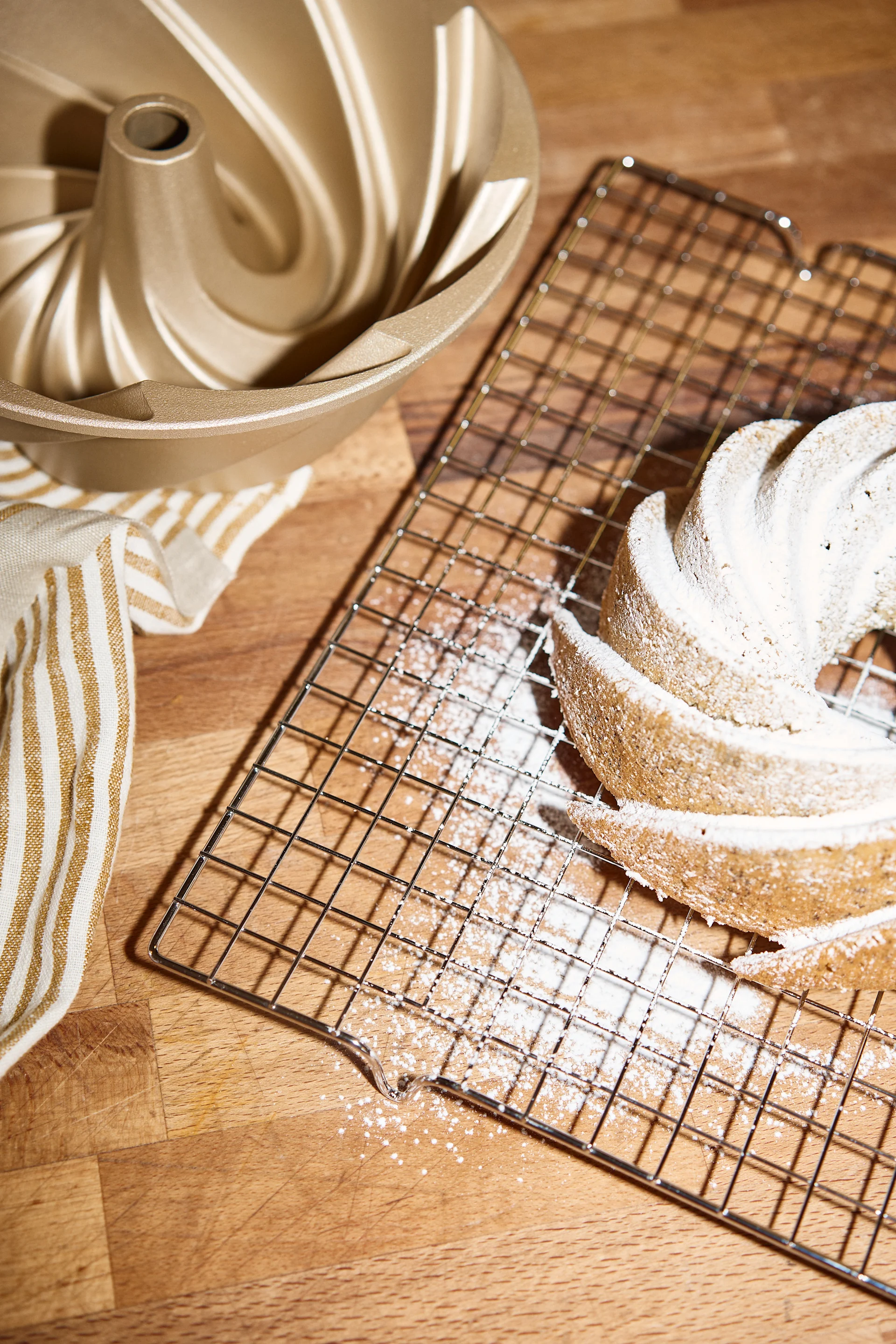 Un moule à bundt doré, une serviette rayée et un gâteau bundt saupoudré de sucre glace sur une grille de refroidissement.