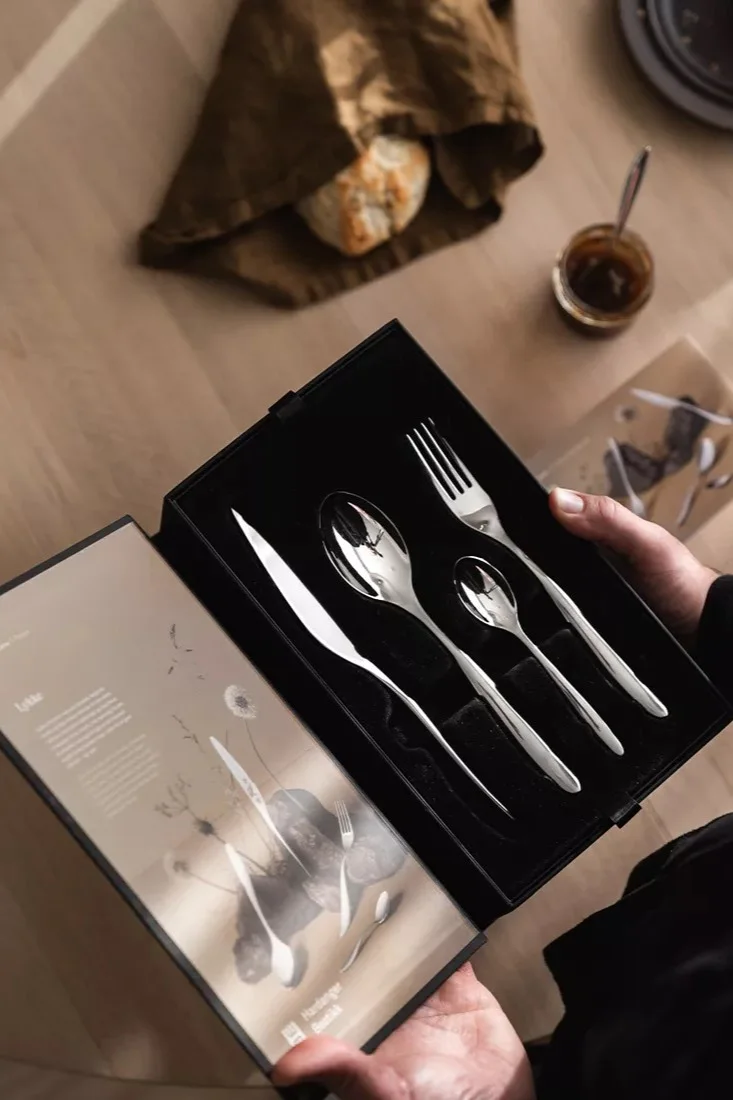 Mains tenant une boîte noire ouverte avec un service de couverts en argent brillant : couteau, grande cuillère, petite cuillère et fourchette. Pain et confiture sur une table en bois.