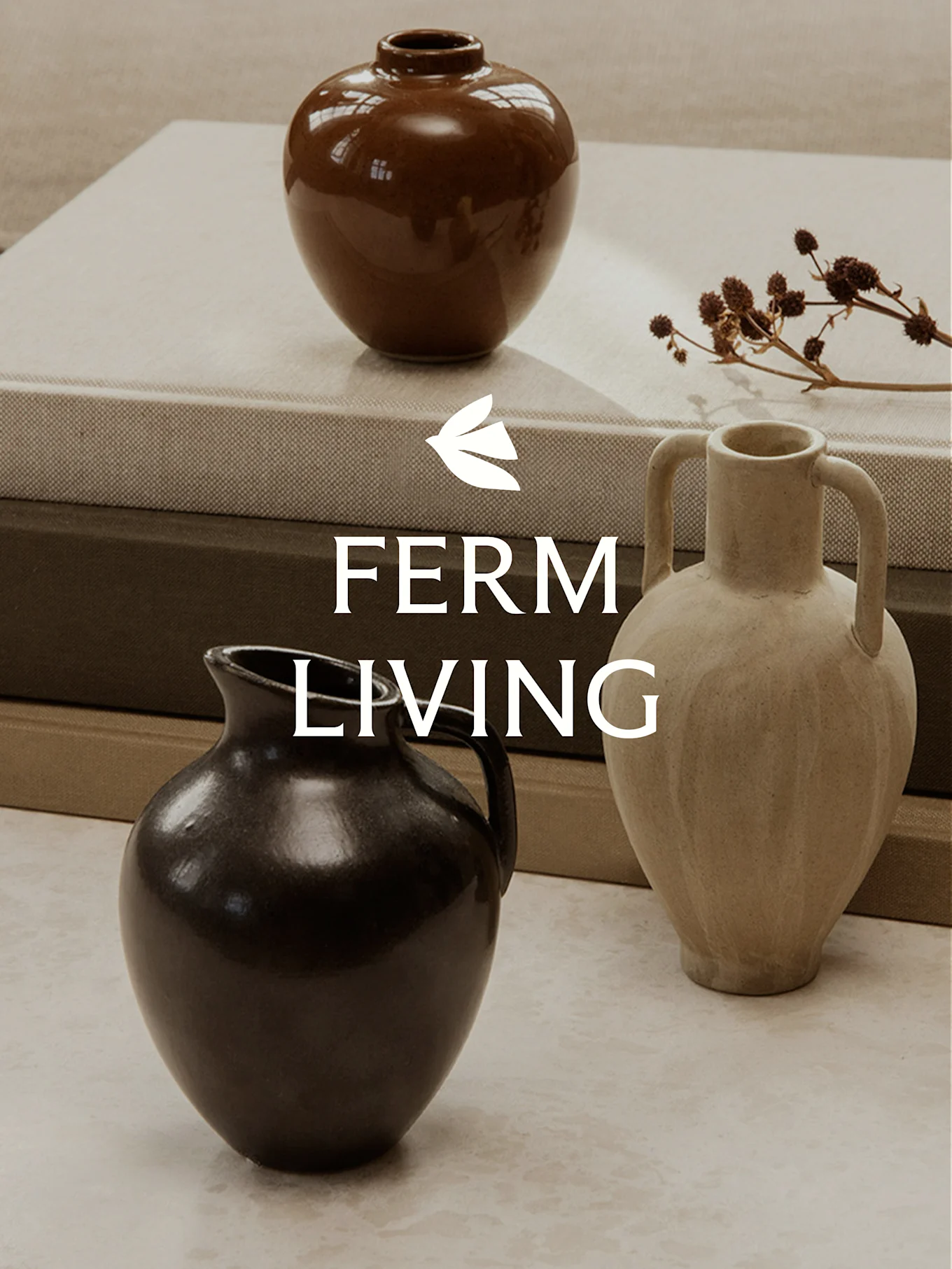 Trois vases en céramique de formes et de couleurs différentes sur des livres empilés, avec des tiges de plantes séchées.