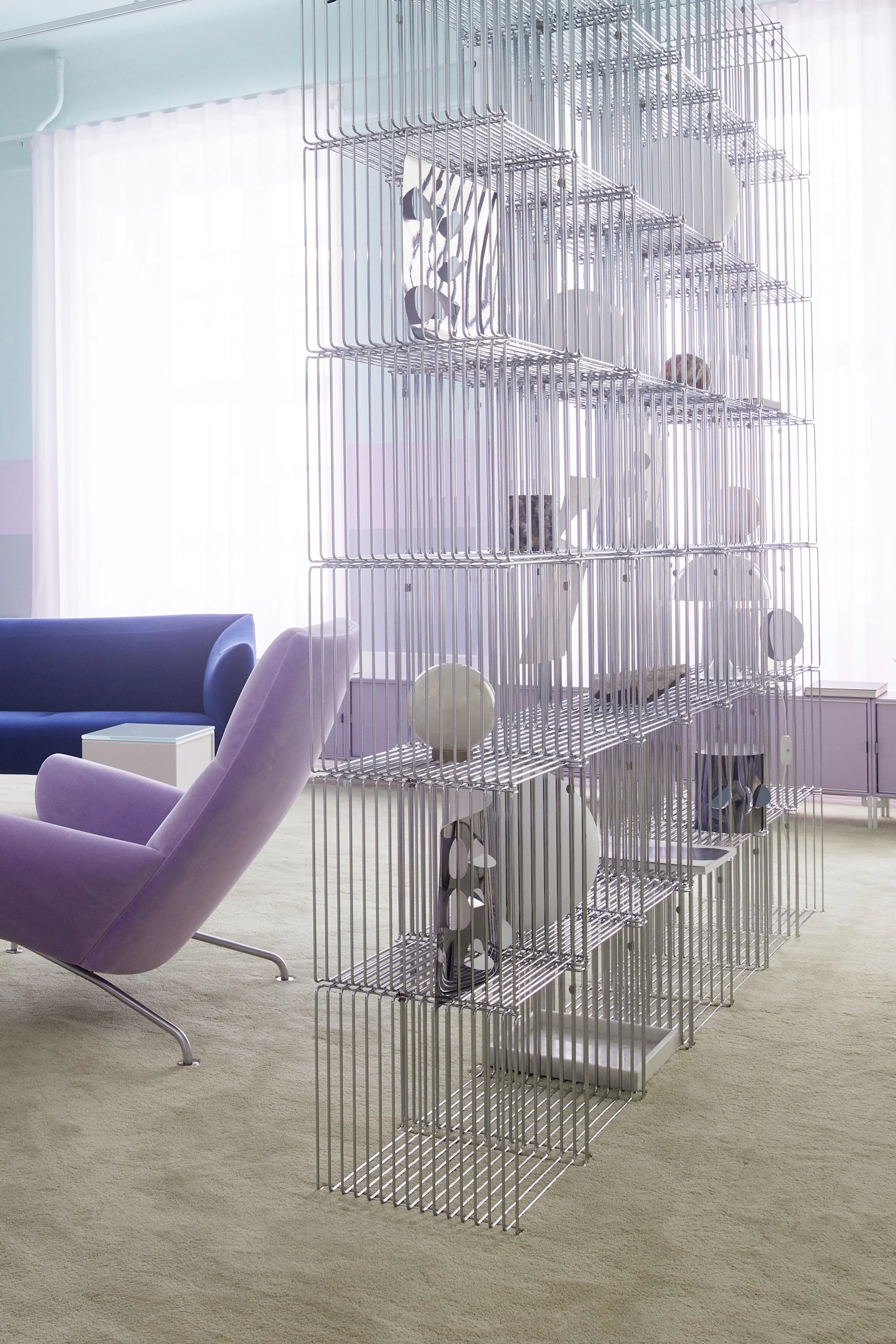 Une pièce moderne avec une haute étagère en fil métallique argenté comme séparateur, un fauteuil violet et un canapé bleu.