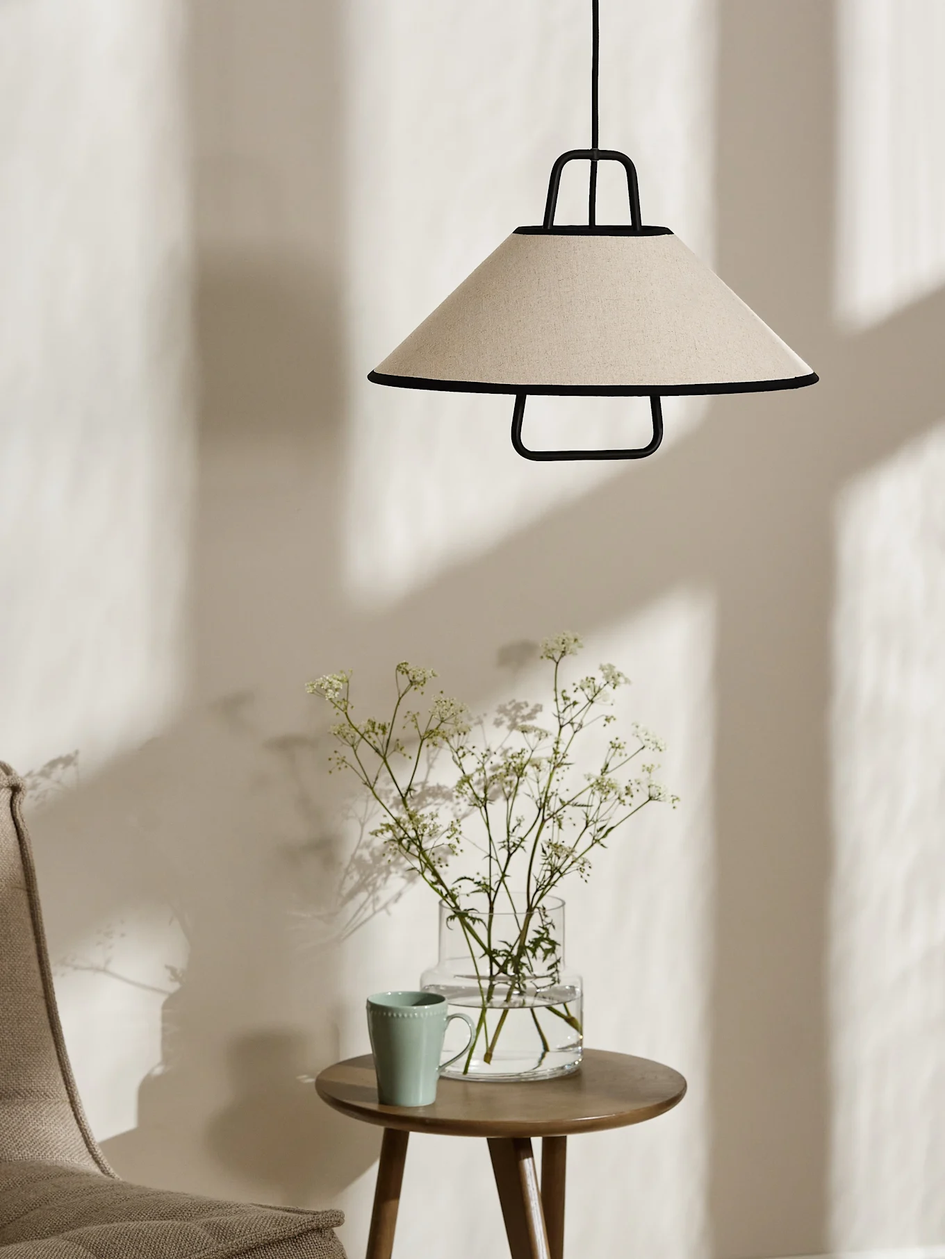 Une lampe suspendue conique de couleur crème avec une bordure noire est suspendue au-dessus d'une table d'appoint en bois avec un vase de fleurs blanches et une tasse vert clair.