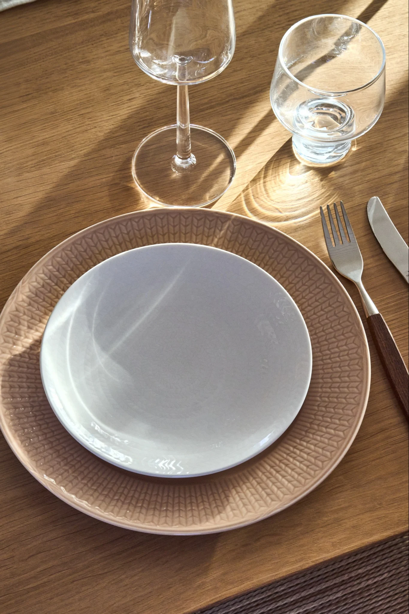 Une table dressée avec une assiette claire sur une assiette beige à motifs, une fourchette, un couteau à manche en bois et deux verres sur une table en bois.
