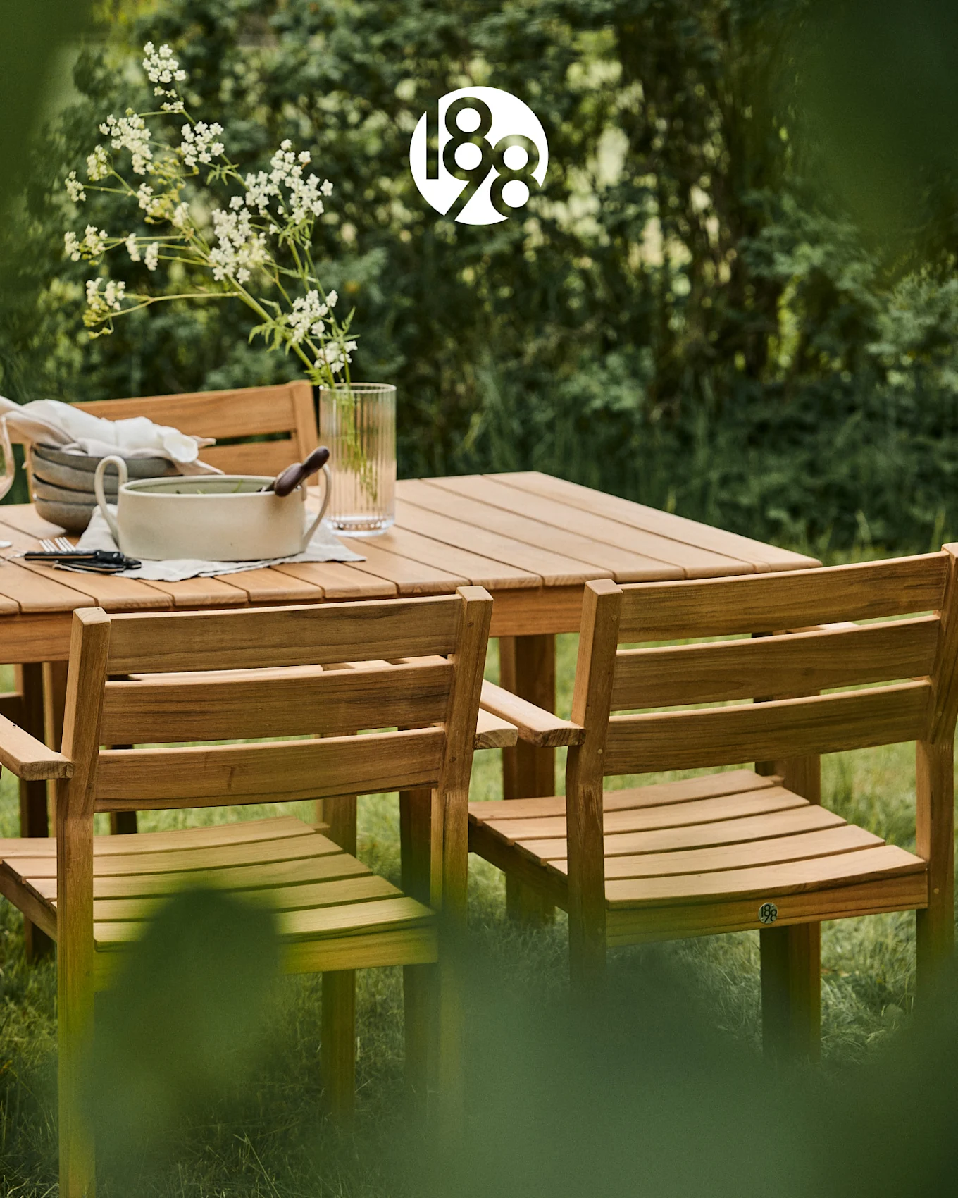 Ensemble de salle à manger extérieur en bois avec table et chaises dans un jardin verdoyant, avec des fleurs blanches et de la vaisselle.