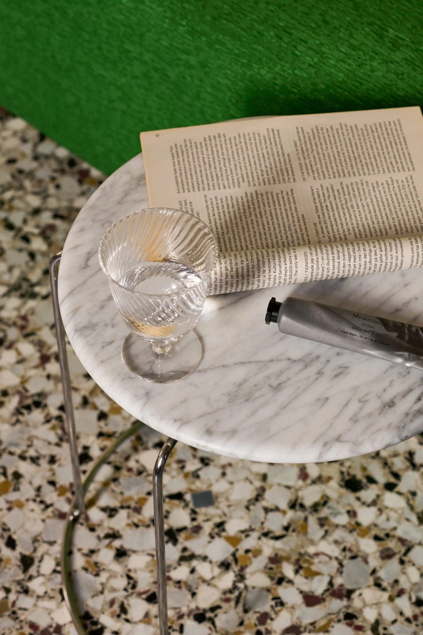 Un verre d'eau transparent strié, un livre ouvert et un tube de baume pour les mains argenté sur une table en marbre.