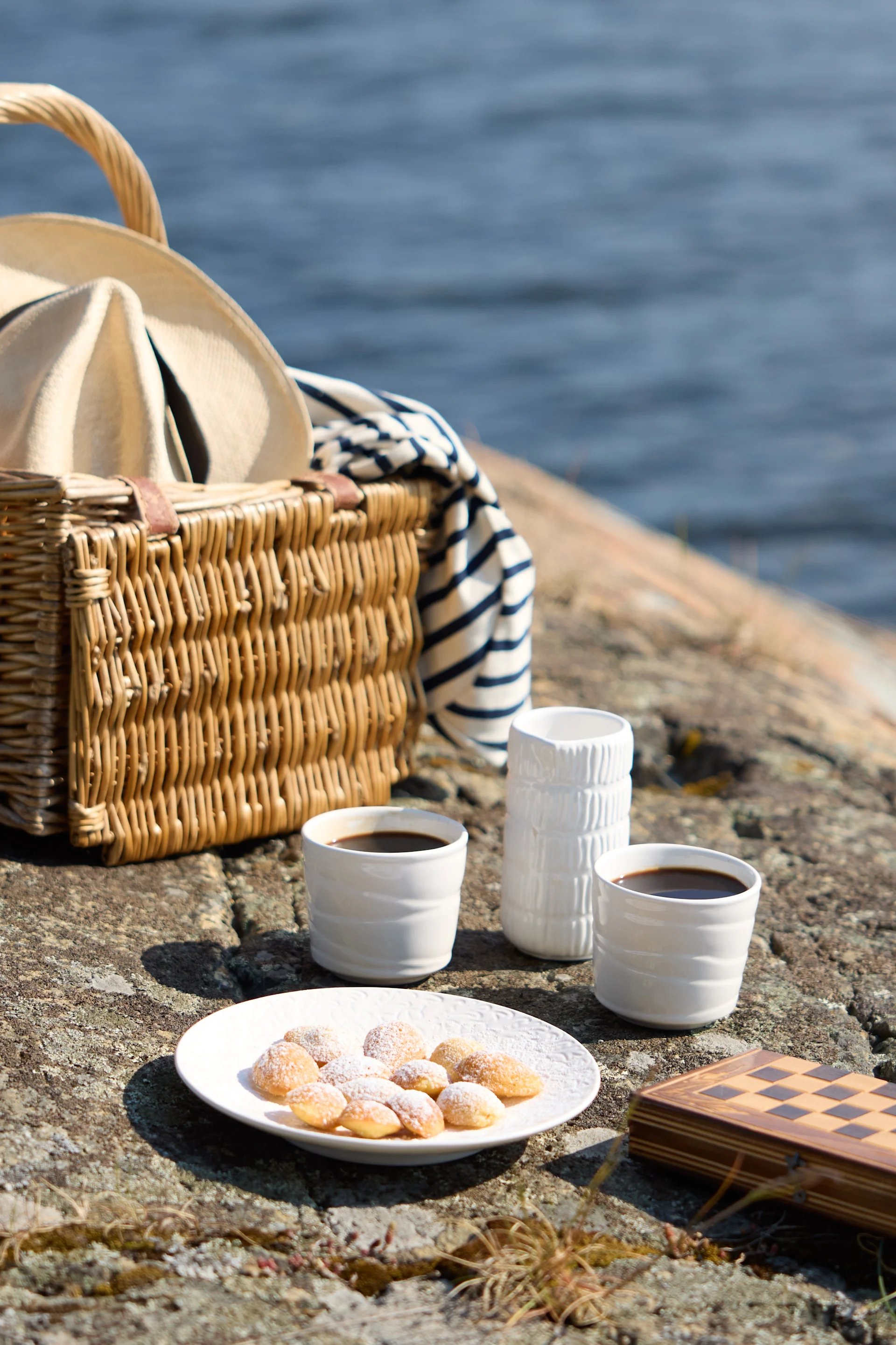 Un pique-nique sur des rochers au bord de la mer, avec un panier en osier contenant un chapeau et une couverture rayée, deux tasses de café, des pâtisseries et un échiquier fermé.