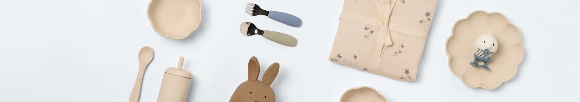 Articles de bébé sur blanc: deux bols crème, couverts bleus et verts, tissu floral, jouet lapin, cuillère en bois, tasse beige et figurine sur assiette festonnée.