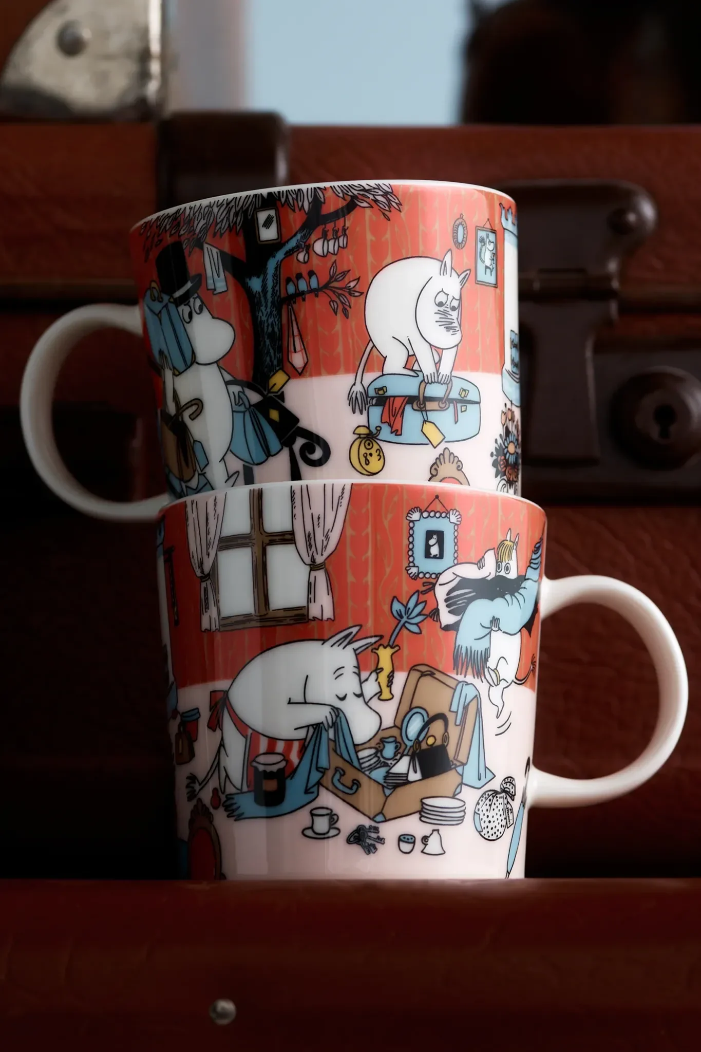 Deux tasses Moomin rouges empilées avec des illustrations de personnages préparant des valises et accrochant des objets à un arbre.