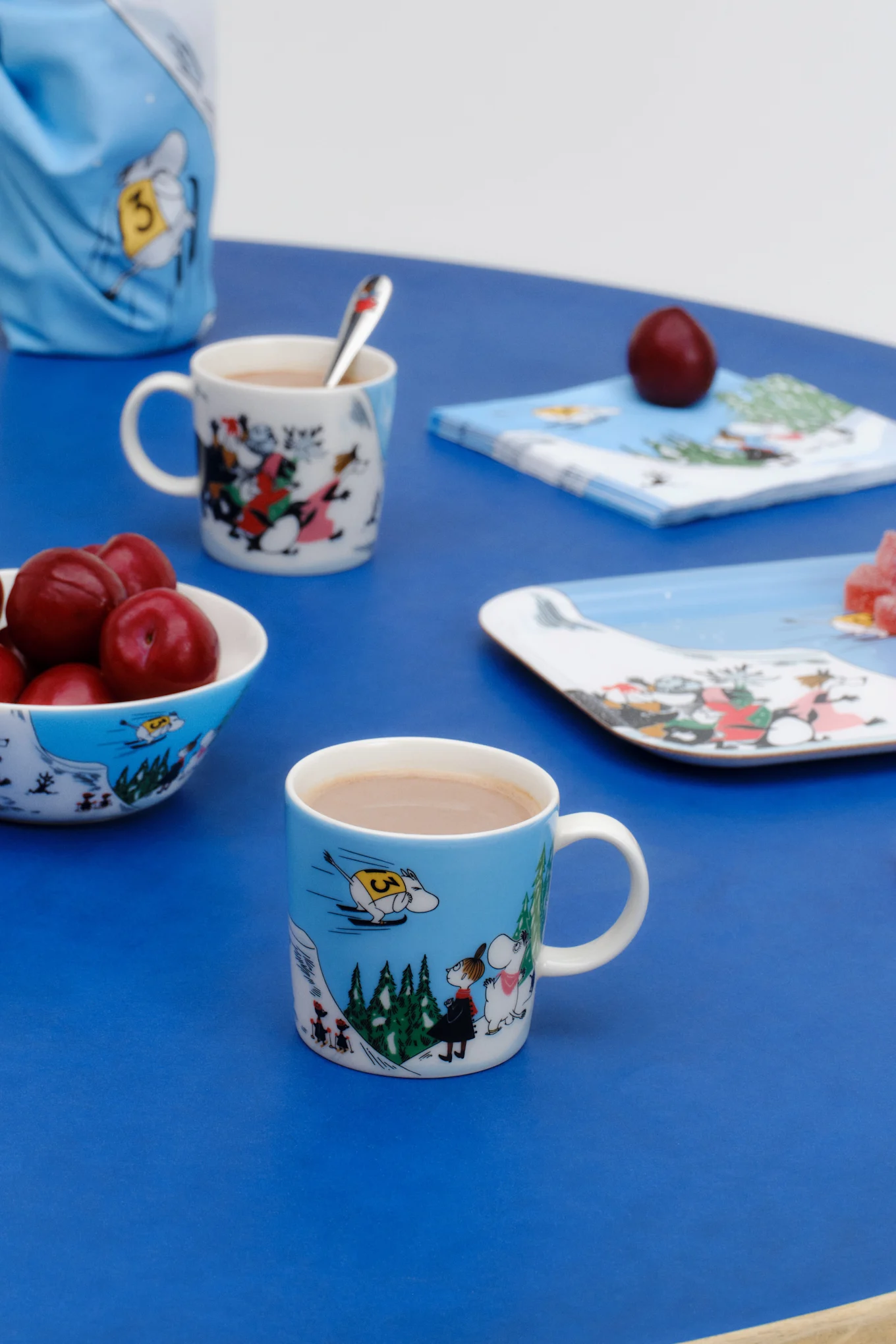 Le mug Moomin Hiver 2024, « Saut à ski », est posé sur une table bleue.