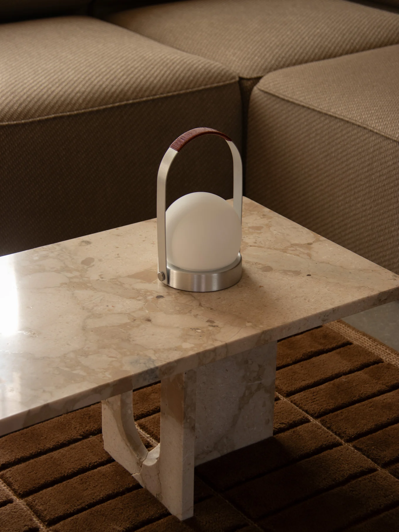 Lampe portable moderne avec lumière blanche sphérique, cadre argenté et poignée en cuir marron sur une table en marbre à côté d'un canapé.