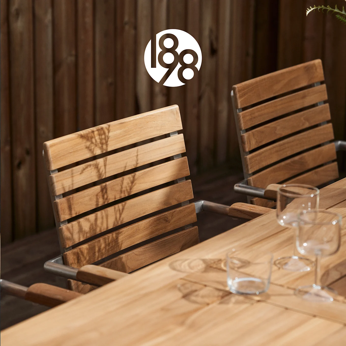 Table et deux chaises de salle à manger en bois extérieur, avec des verres, sous un logo 188, baignées de soleil.