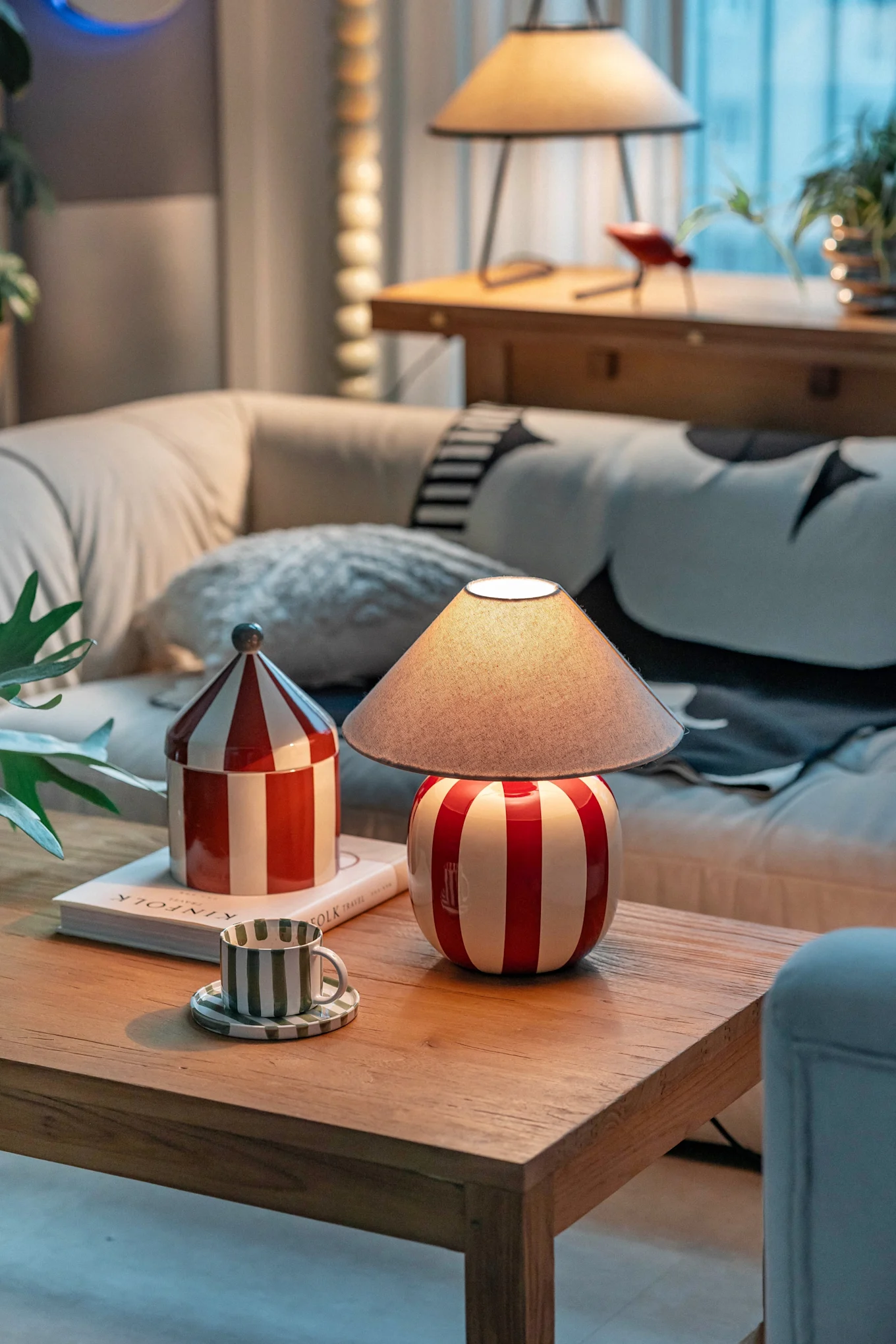 Un salon confortable avec une lampe à rayures rouges et blanches, un pot assorti et une tasse à rayures vertes sur une table basse en bois.