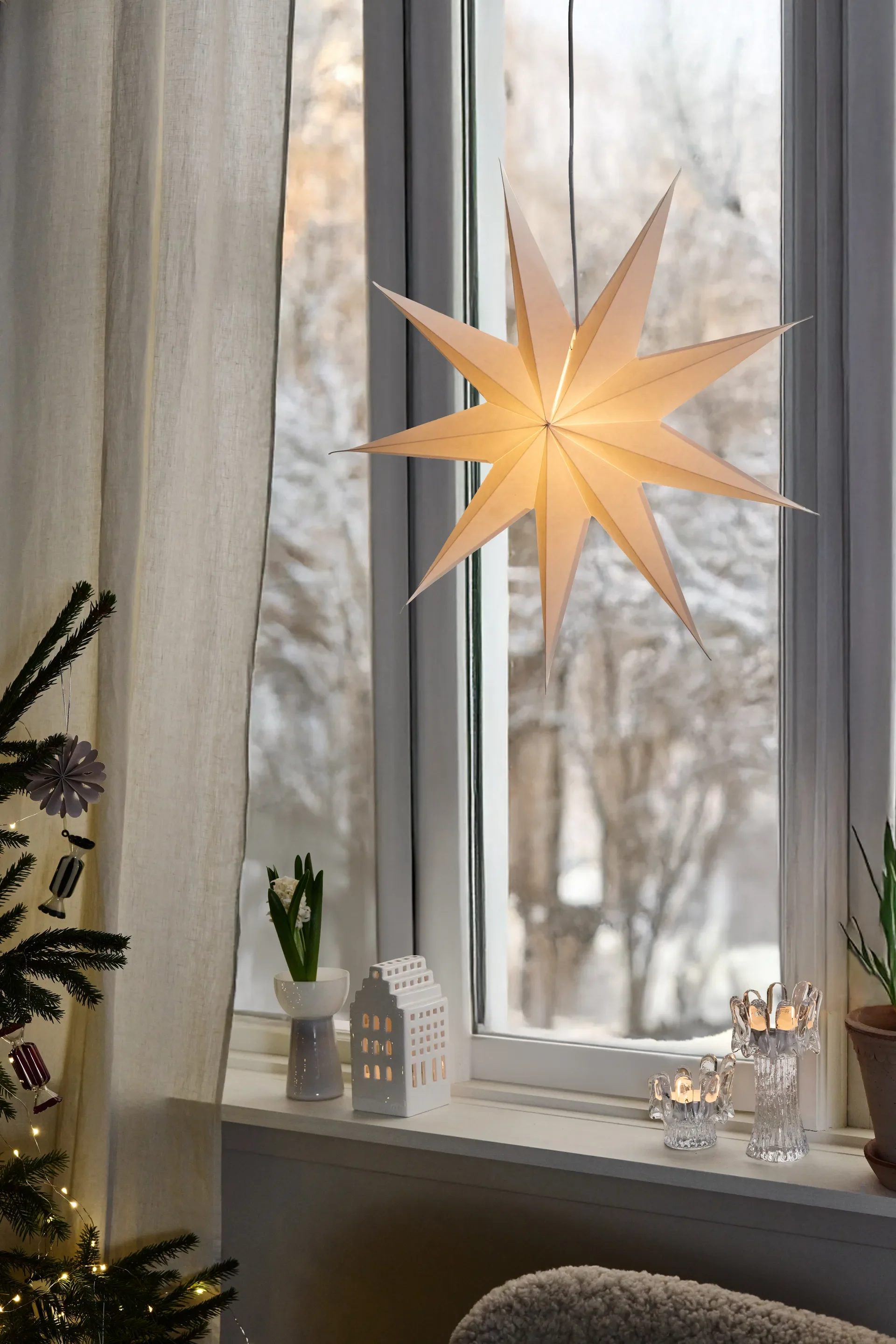 Fenêtre hivernale décorée: étoile en papier lumineuse, maison en céramique, photophores et sapin de Noël.