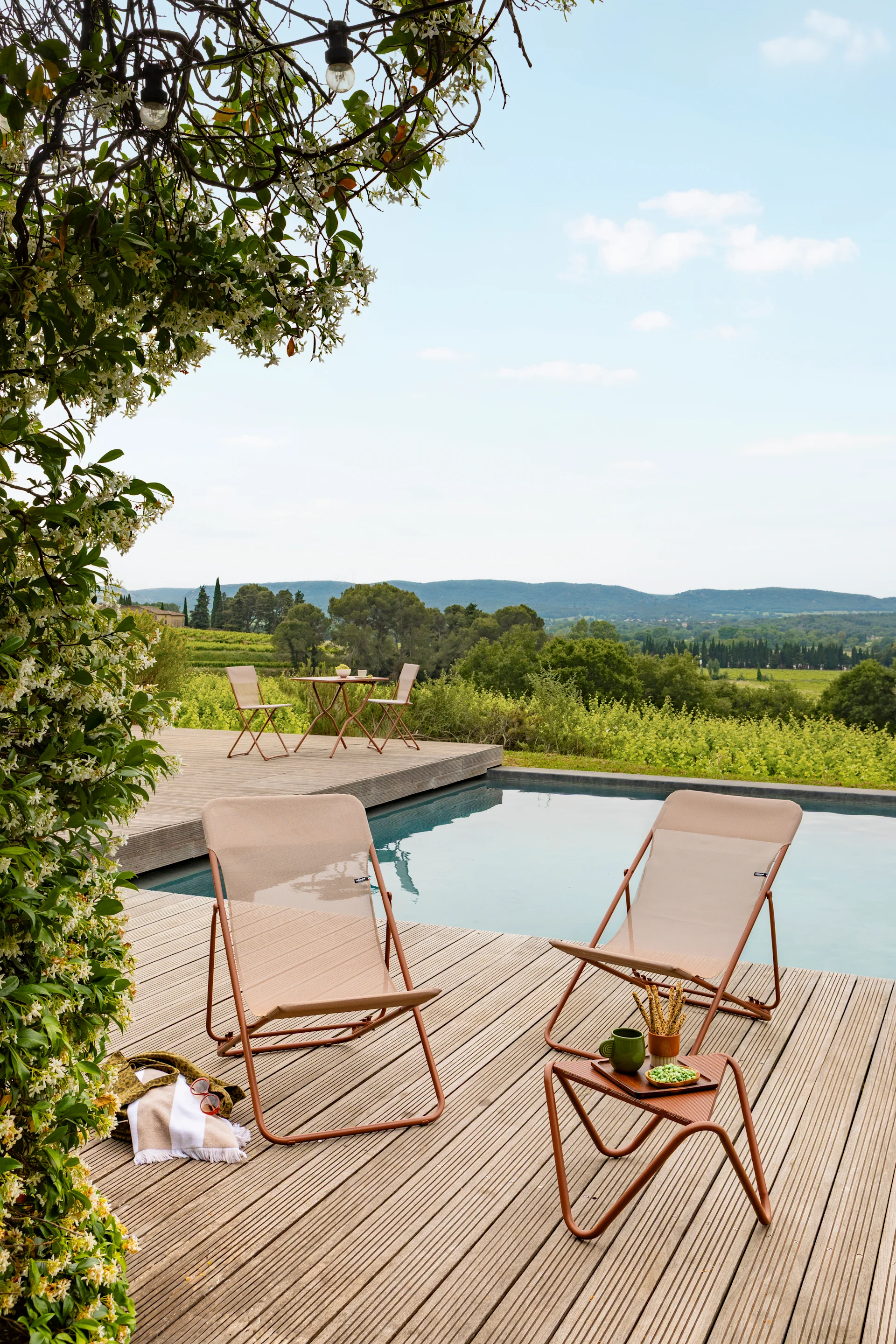 Deux chaises longues d'extérieur beige et une table d'appoint sur une terrasse en bois près d'une piscine, avec des vignes fleuries et vue sur un vignoble.