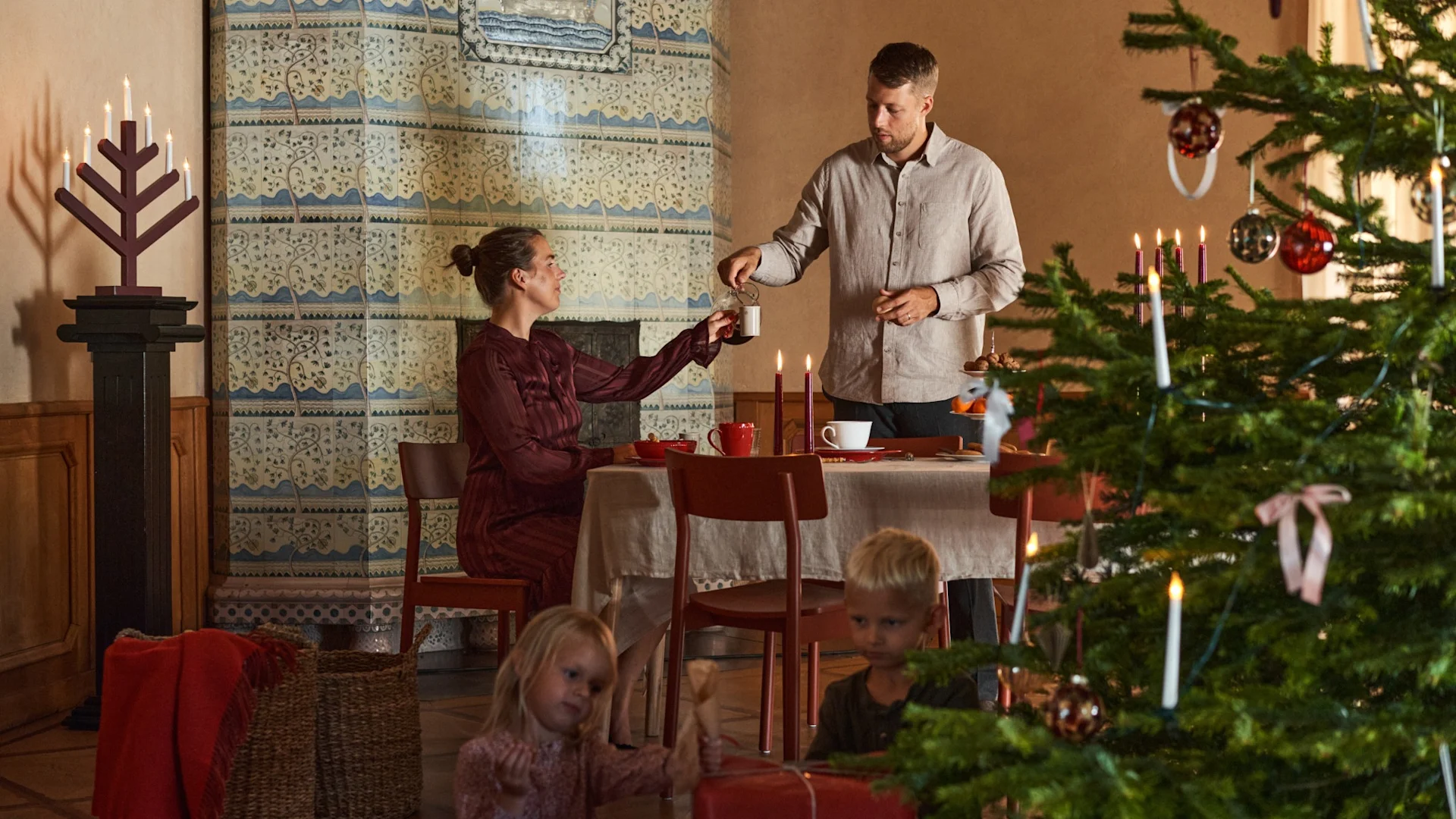 Un couple est assis à une table décorée pour Noël, à côté d'un sapin de Noël avec des enfants qui déballent leurs cadeaux.