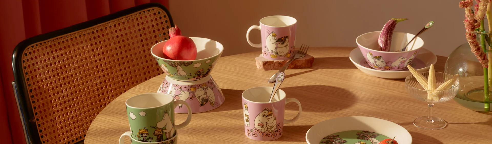Une table en bois dressée avec des tasses, bols et assiettes Moomin verts et roses, une grenade, une aubergine et du maïs bébé.
