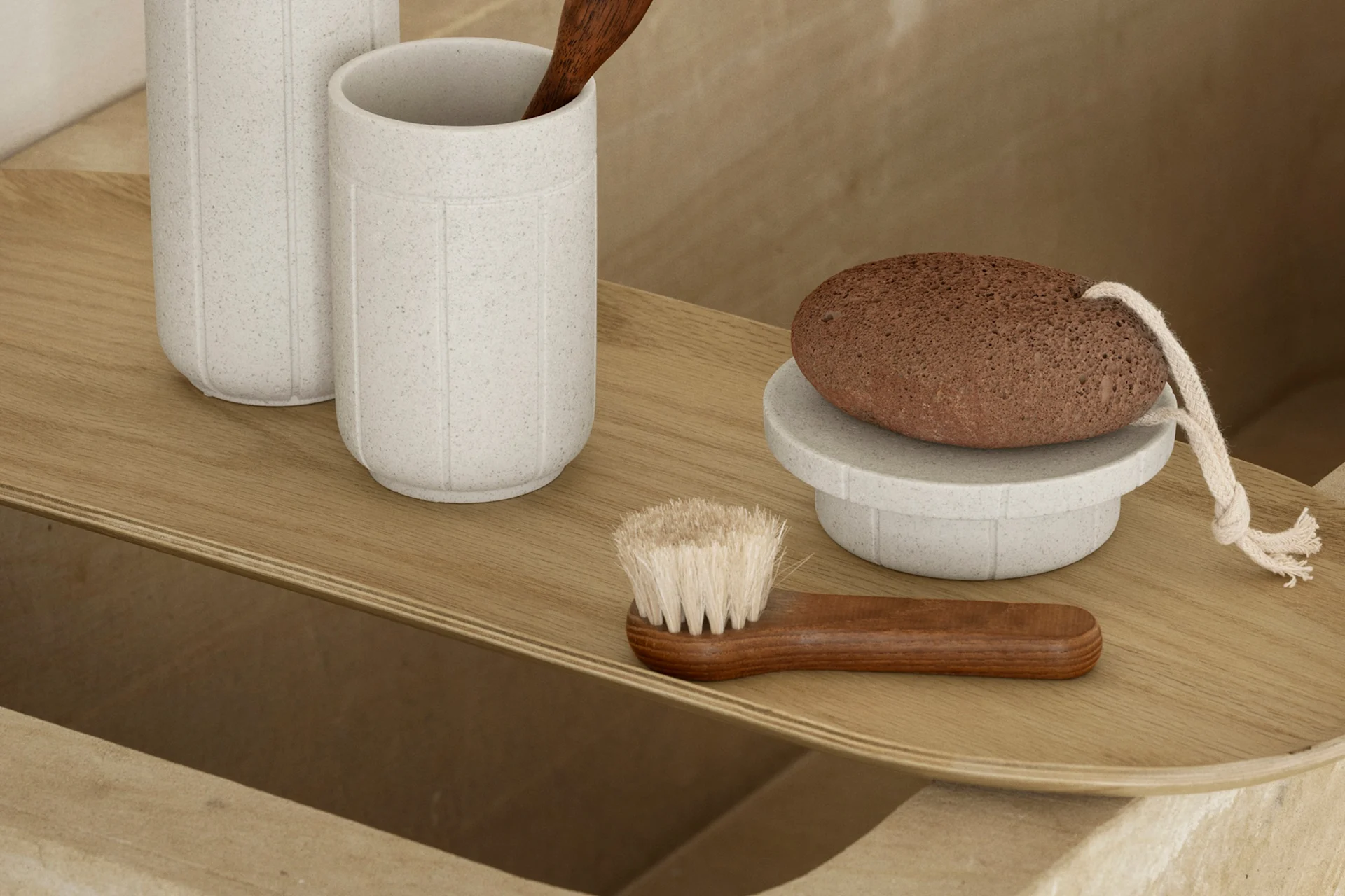Un plateau en bois clair avec des accessoires de bain : deux tasses grises mouchetées, une cuillère en bois, une pierre ponce brune sur un support et une brosse en bois.