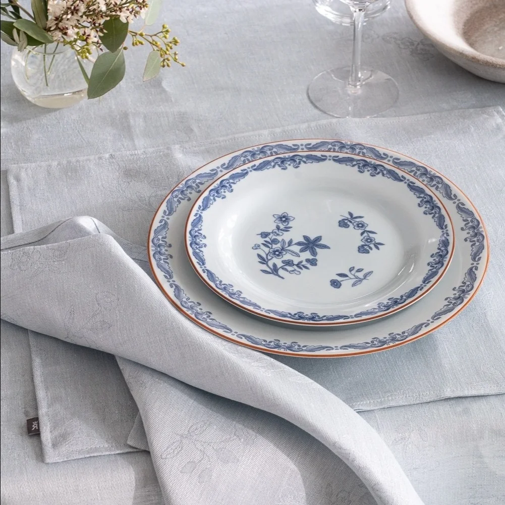 Assiettes blanches empilées avec motifs floraux et ornementaux bleus, set de table bleu clair et serviette pliée sur une table.
