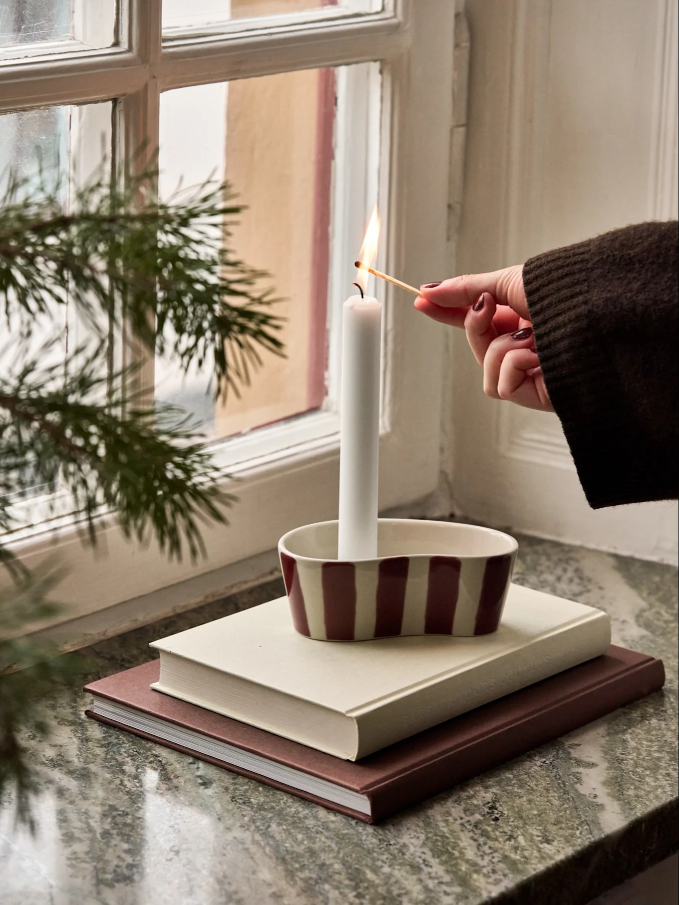 Une main allume une bougie blanche dans un support rayé sur une pile de livres près d'une fenêtre avec des branches de pin.