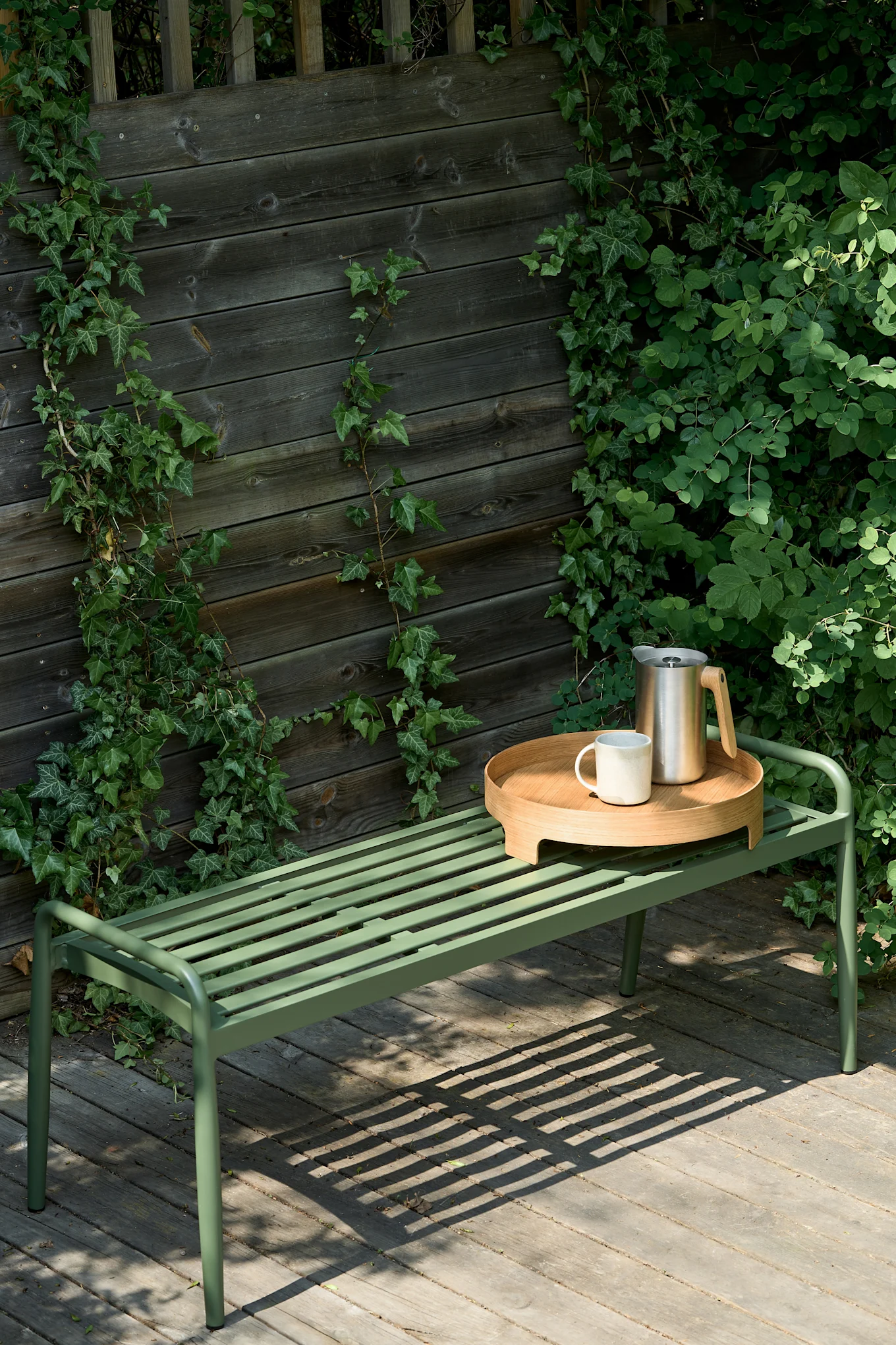 Banc métallique vert à lattes sur terrasse en bois, avec plateau, tasse blanche et carafe en inox, près d'une clôture de lierre.
