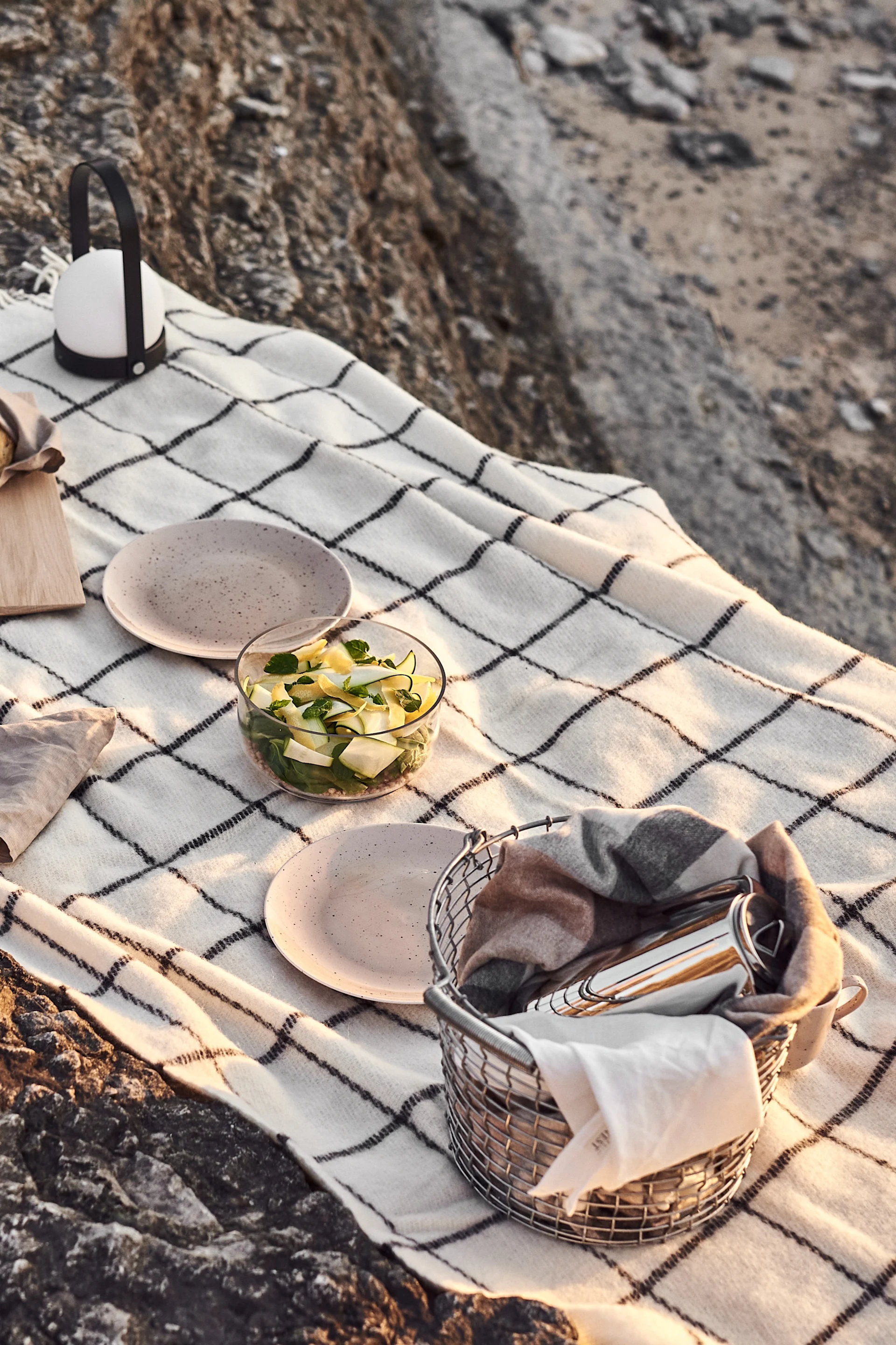 Une couverture de pique-nique posée sur des rochers, avec une salade, des assiettes, une lanterne et un panier contenant des couverts.