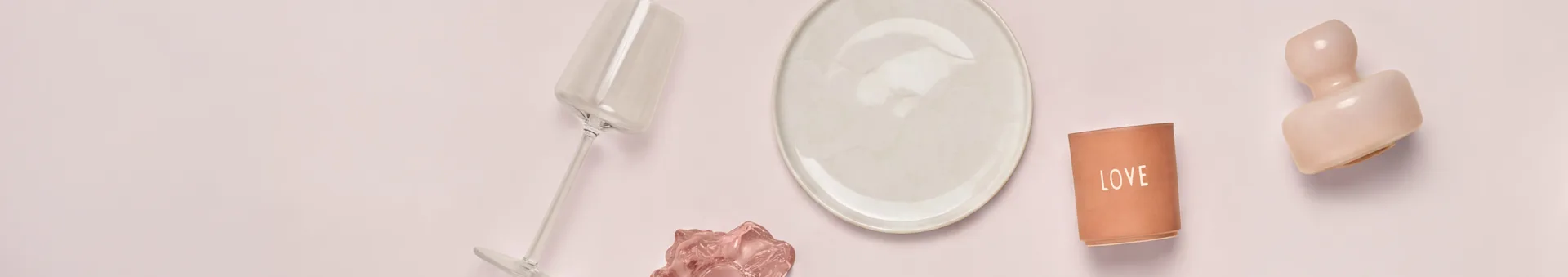 Vue de dessus de divers objets décoratifs et de table sur une surface rose clair. De gauche à droite : un verre transparent incliné, une assiette crème, un bougeoir en terre cuite "LOVE" et une sculpture abstraite rose. Un objet ondulé rose plus petit est sous le verre.