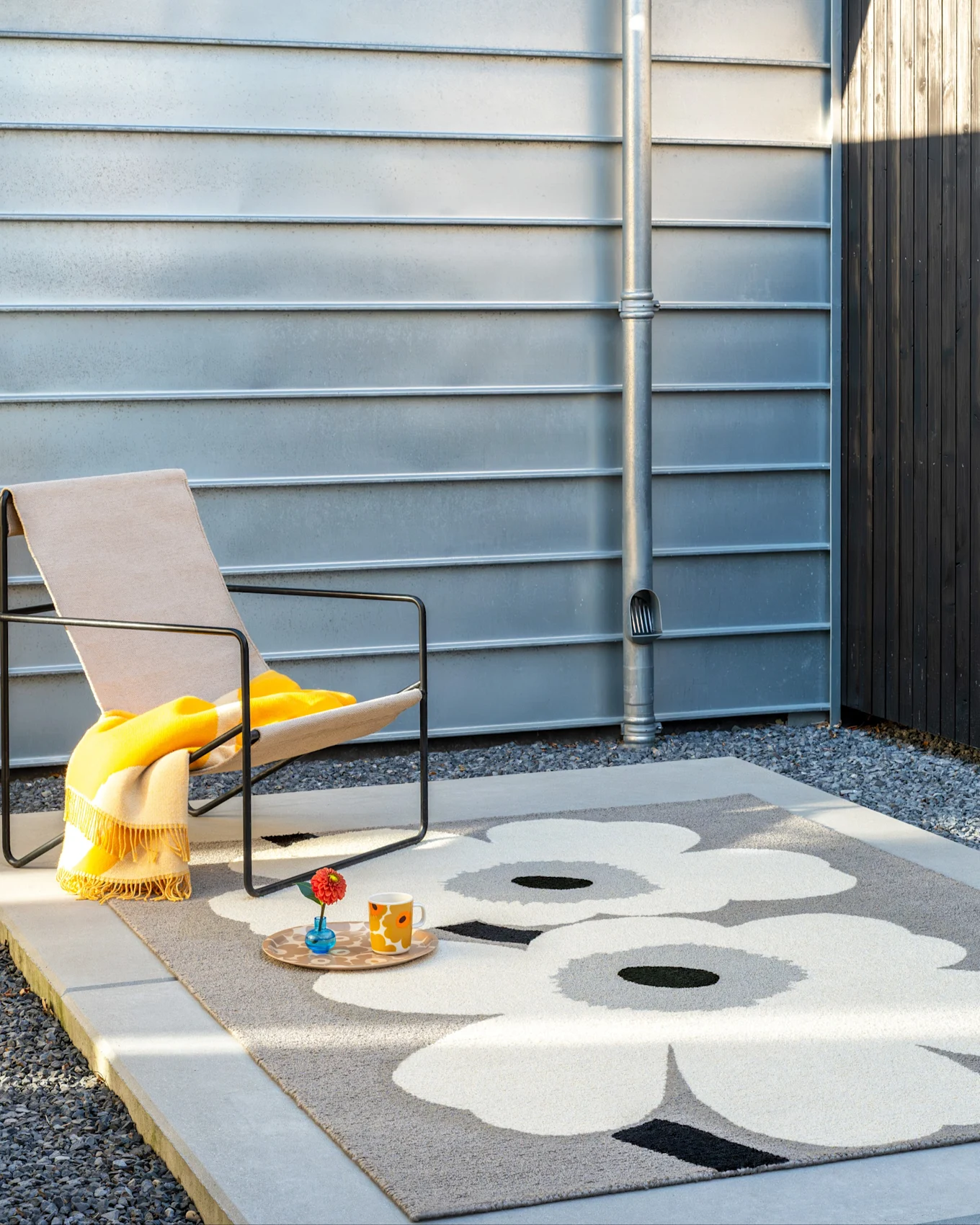 Scène extérieure: chien sur tapis gris à fleurs blanches, près d'une chaise avec couverture jaune et mur métallique.