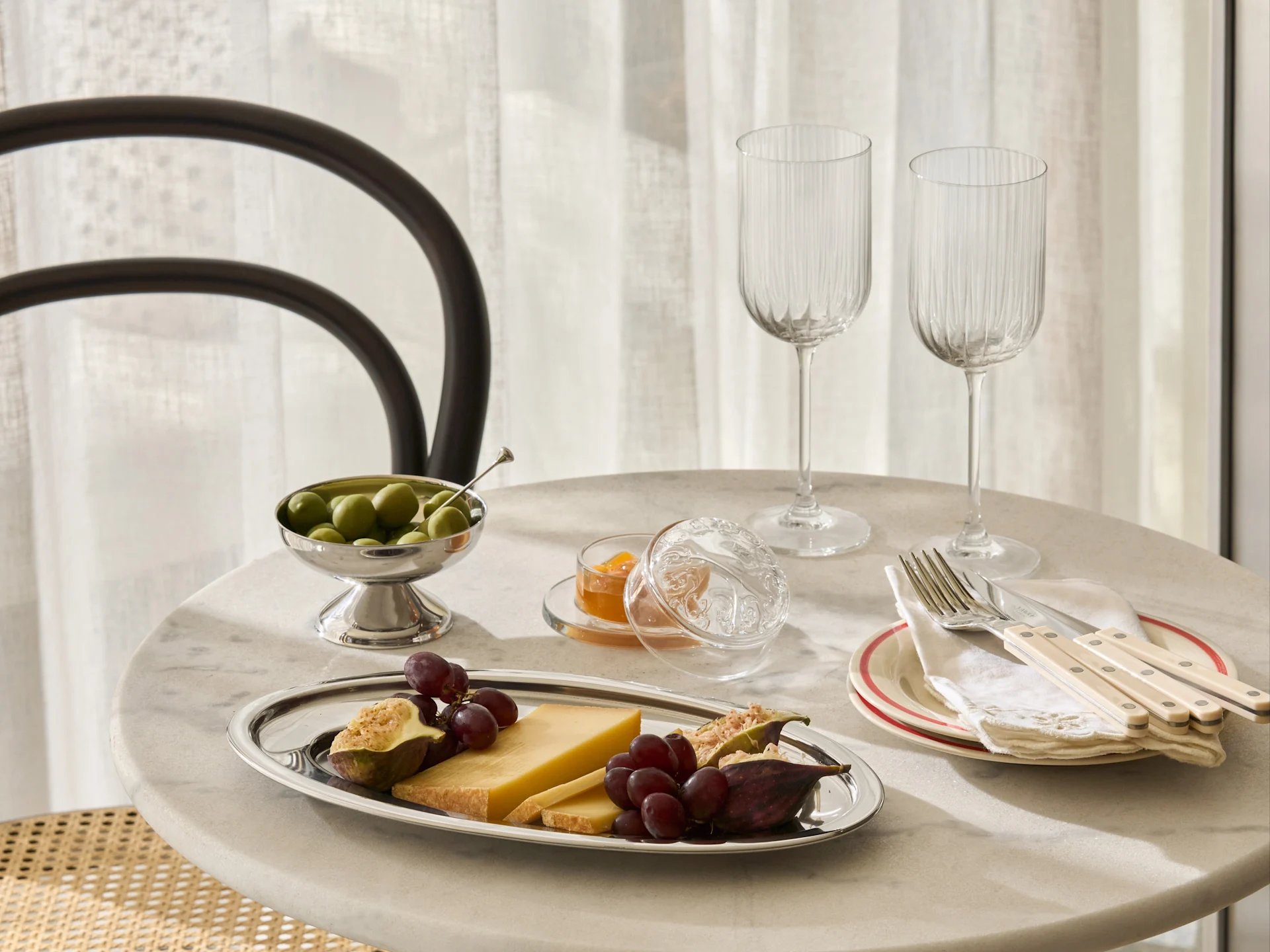 Une table en marbre dressée avec un plateau argenté de fromage, raisins, figues, un bol d'olives, de la confiture, deux verres nervurés et des couverts sur des assiettes.