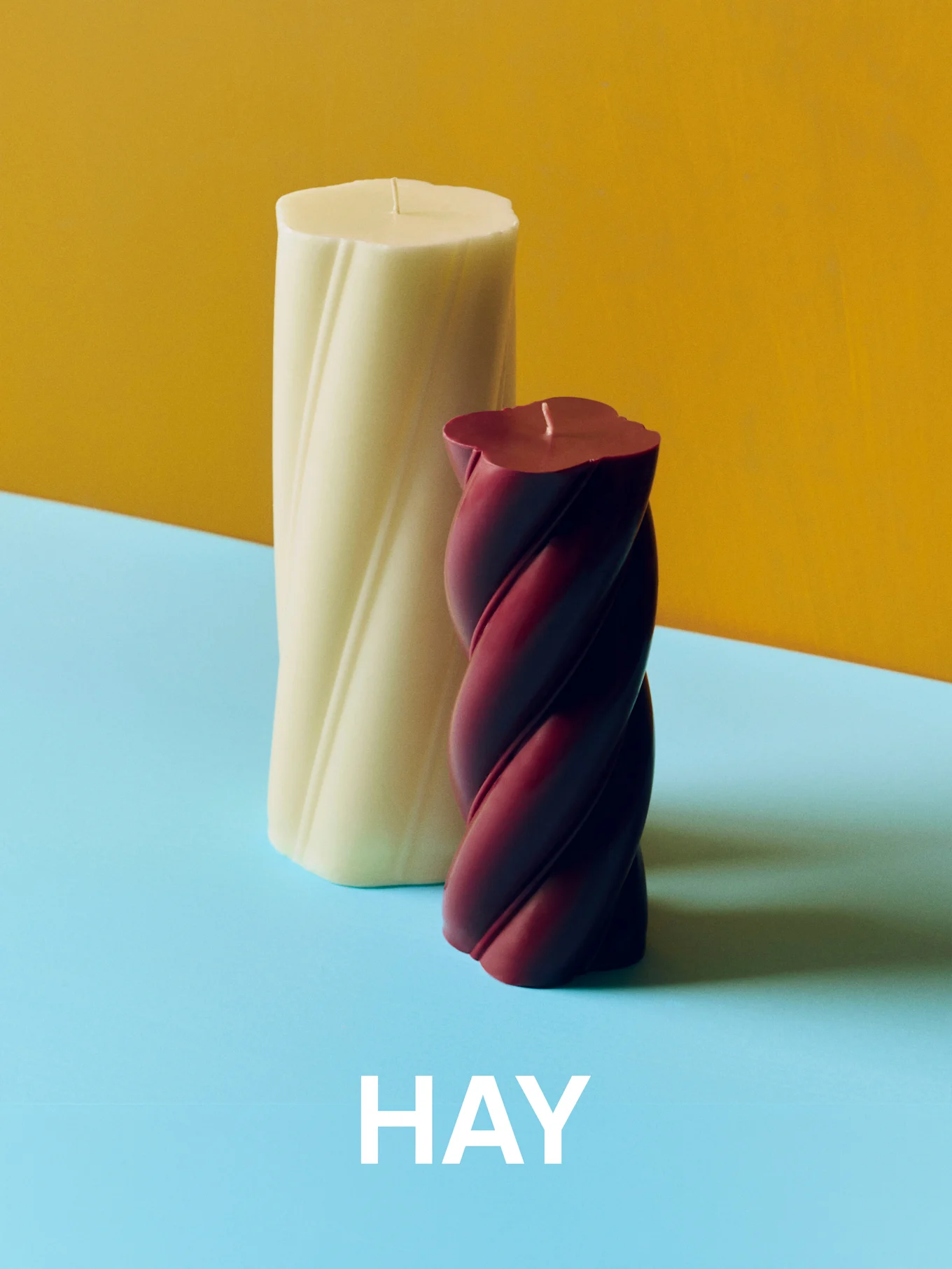 Bougies torsadées crème et bordeaux sur fond jaune et bleu avec le texte 'HAY'.