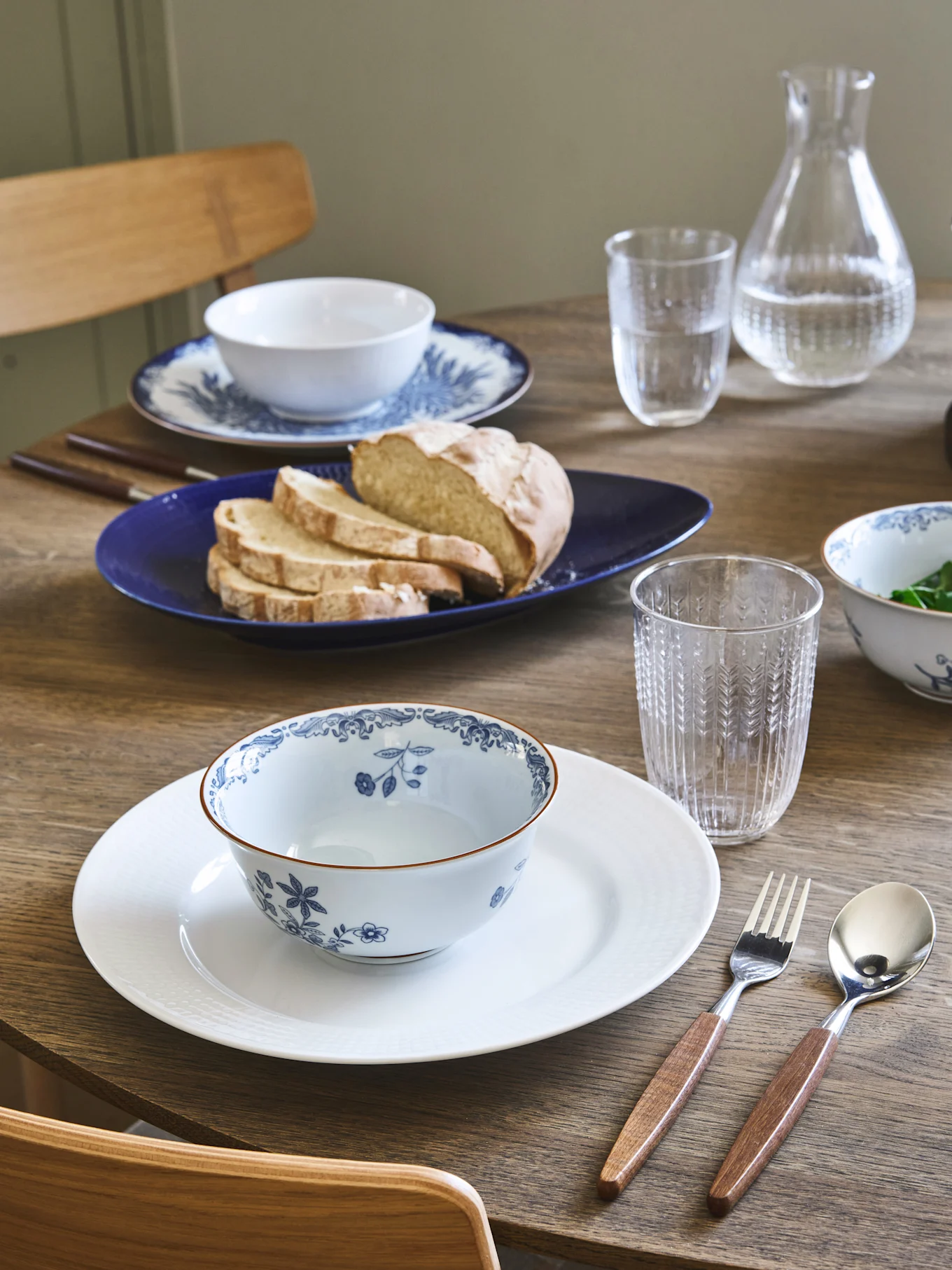 Table à manger dressée avec deux couverts, bols fleuris bleus et blancs, du pain sur une assiette bleue et des couverts à manche en bois.