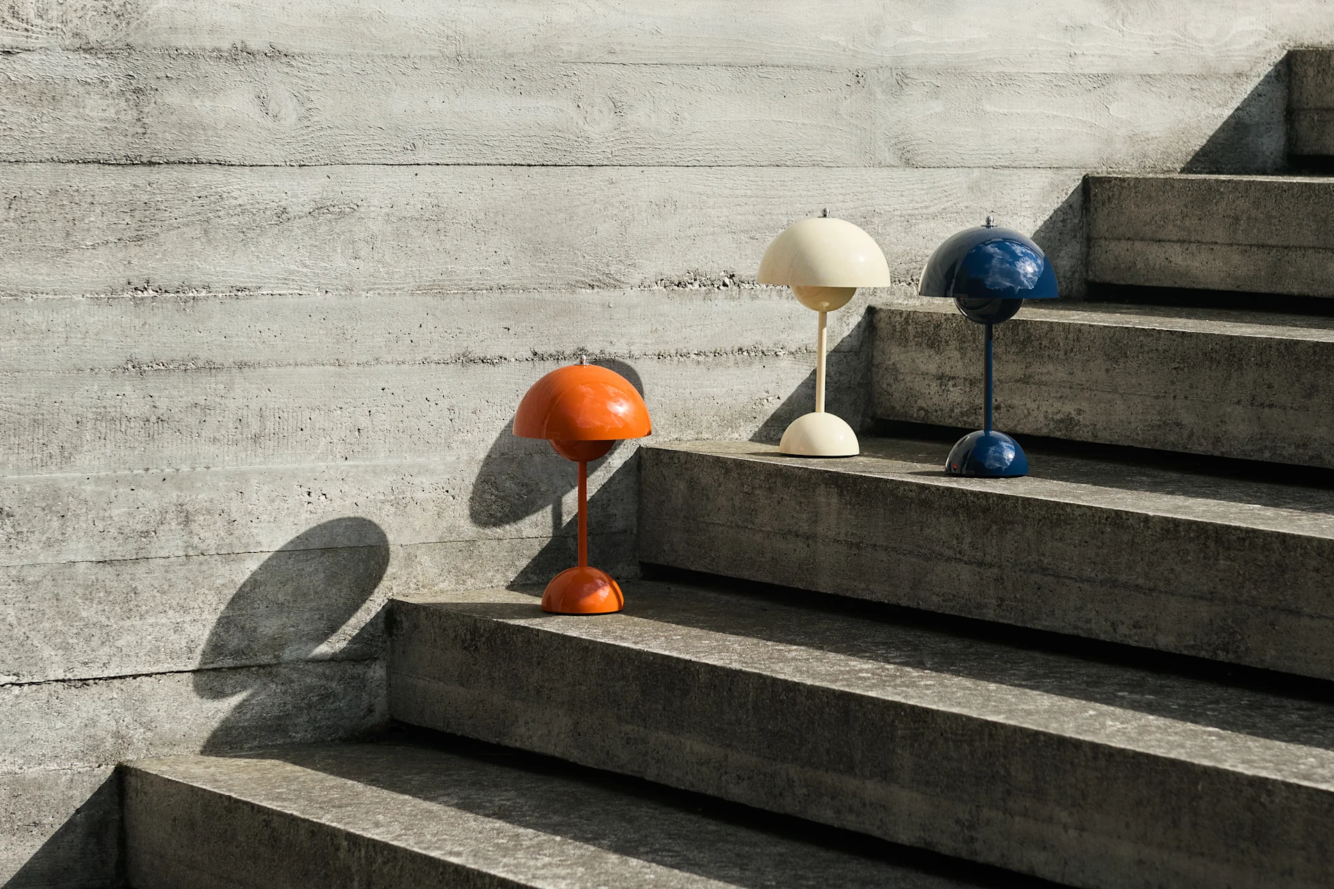 Trois lampes de table en forme de dôme, orange, crème et bleu, sur des escaliers en béton près d'un mur en béton, projetant des ombres.