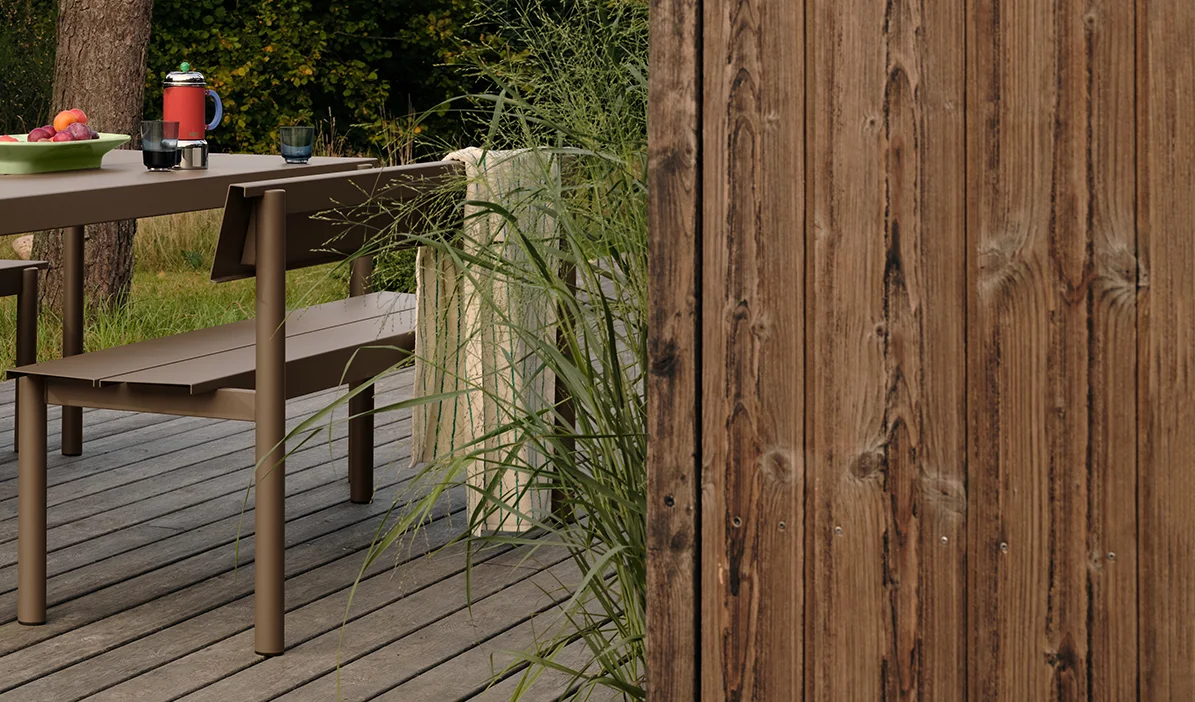 Table et banc d'extérieur en métal marron sur une terrasse en bois avec fruits et boissons, près d'herbes hautes et d'une clôture en bois.